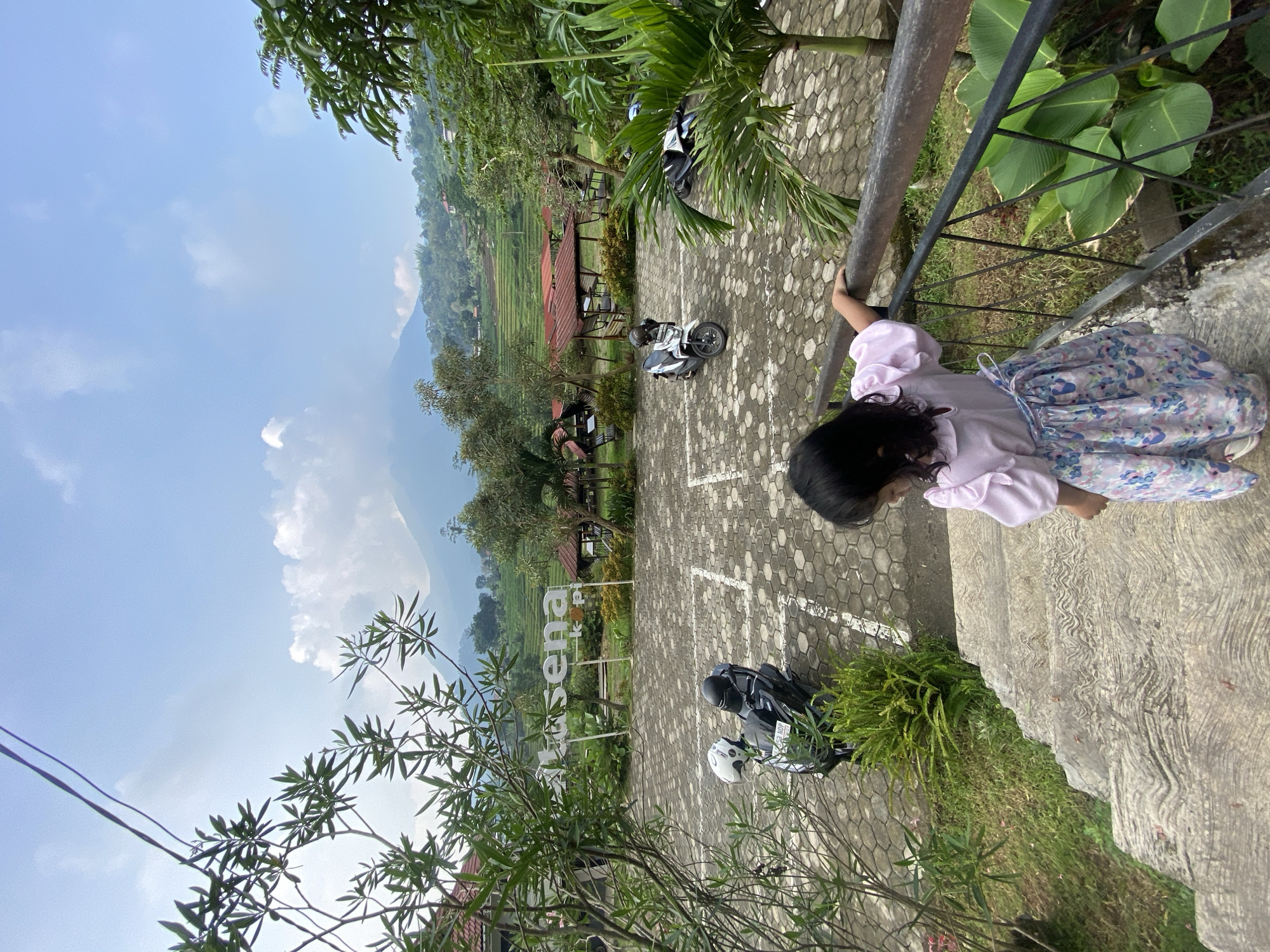 A young girl with dark hair, wearing a light pink shirt and a light blue floral skirt, stands on a concrete platform and leans against a metal railing, looking down. The location appears to be a scenic overlook or viewing area in Penanggungan, Indonesia. The ground is paved with hexagonal concrete blocks, marked with white painted parking spaces. Several parked motorcycles are visible in the parking area. Beyond the parking area, there is a view of rice paddies, trees, and mountains. There are also small structures with red roofs visible amongst the trees. The sky is blue with scattered clouds. Power lines are visible in the foreground. Vegetation grows along the edge of the platform where the girl is standing.