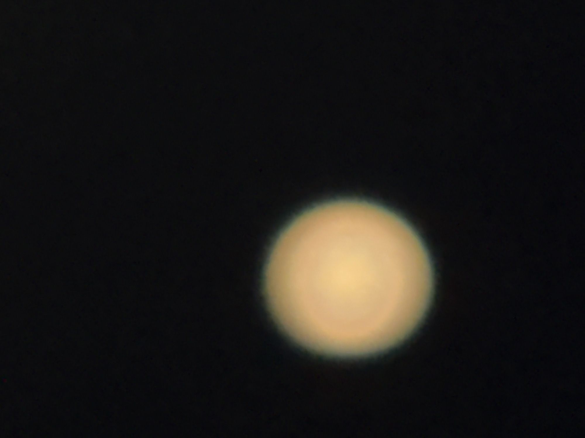 The image shows a faint, out-of-focus circular object against a dark background. The object appears to be a celestial body, possibly a planet or moon, with a soft, yellowish-orange hue and a slightly hazy edge. Concentric rings of lighter and darker shades are subtly visible within the object, suggesting some surface detail or atmospheric banding, though the blurriness of the image makes definitive identification difficult. The background is a uniform, deep black, indicative of an observation in space or a very dark environment. There is no text visible in the image. The context of Mikhaylovsk, Russia, suggests this may be an astronomical observation taken from that location, likely at night. The overall impression is one of looking at a distant, luminous sphere in the vastness of space.