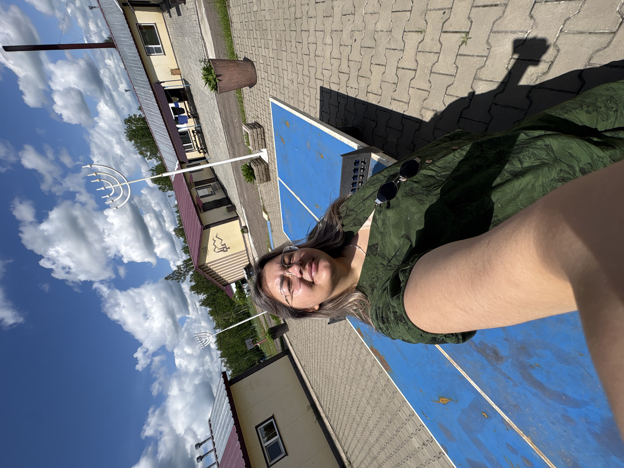 A woman with glasses and a green patterned dress takes a selfie outdoors. She is positioned in front of a blue ping pong table. In the background, there are several low, single-story buildings with red roofs, a white menorah sculpture, trees, and a bright blue sky with white clouds. The ground is paved with interlocking concrete blocks. The location appears to be a recreational area, possibly within or near the city of Birobidzhan, Russia, given the presence of the menorah, a symbol of Jewish culture and heritage.