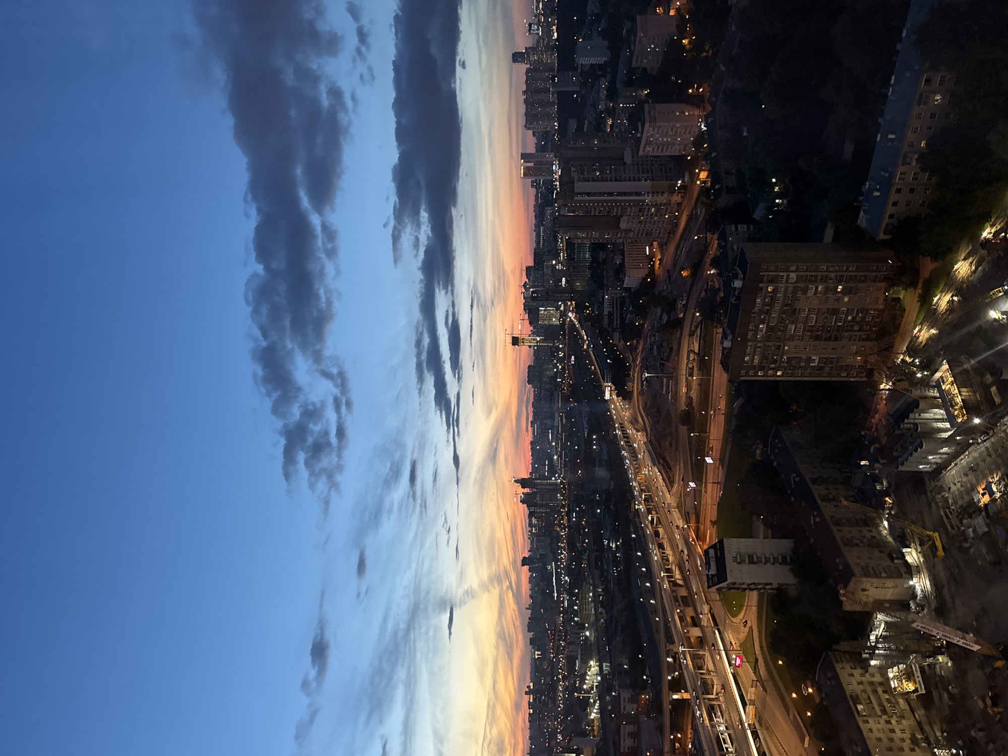 An aerial, wide-angle, vertical shot captures a dusk cityscape. The sky transitions from deep blue at the top to hues of orange, pink, and yellow near the horizon, with scattered clouds casting shadows. Below, a sprawling city unfolds, illuminated by streetlights, building windows, and the headlights of cars on a multi-level highway interchange that dominates the mid-ground. Several high-rise buildings are visible in the distance, their lights twinkling. In the foreground, construction cranes and partially built structures suggest ongoing development. The image appears to be taken from a high vantage point, looking out over a densely populated urban area. This scene is consistent with a view of the city of Khimki, Russia, given its urban landscape and proximity to major transportation routes often seen in aerial photography of the area.