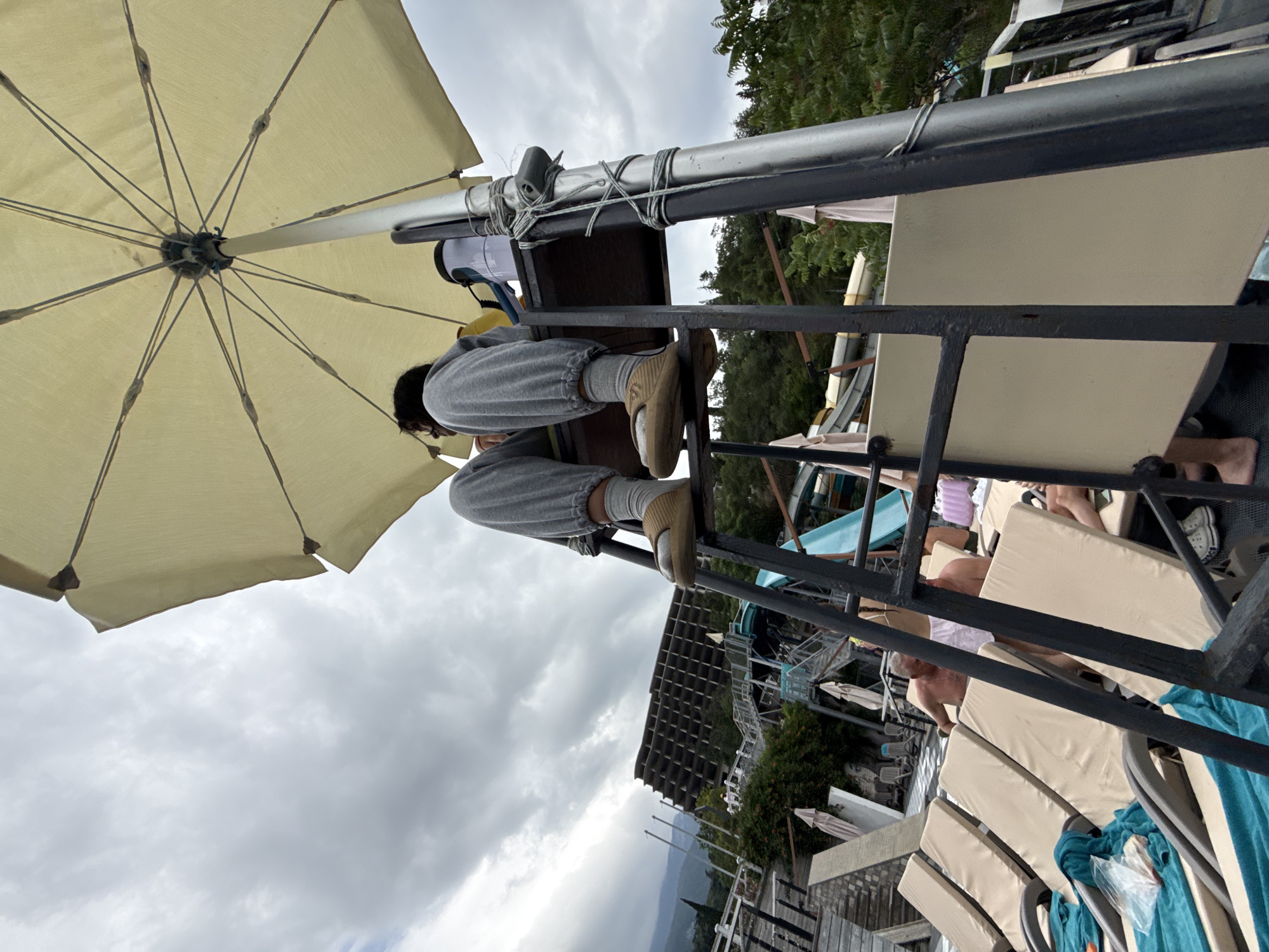 An eye-level, low-angle shot captures a person in gray sweatpants and tan slides sitting on a lifeguard chair under a large cream-colored umbrella. The person is looking to their left, out of frame. The chair has a ladder leading up to it. In the background, there is a large, dilapidated, multi-story building with a grid-like facade, suggesting an abandoned resort or hotel. Below the building, a water park is visible with blue slides and stairs. Several lounge chairs are set up near a pool area. The sky is overcast with clouds. The image appears to be taken in a resort area, potentially in Yalta, Ukraine, given the architectural style and the presence of a water park.