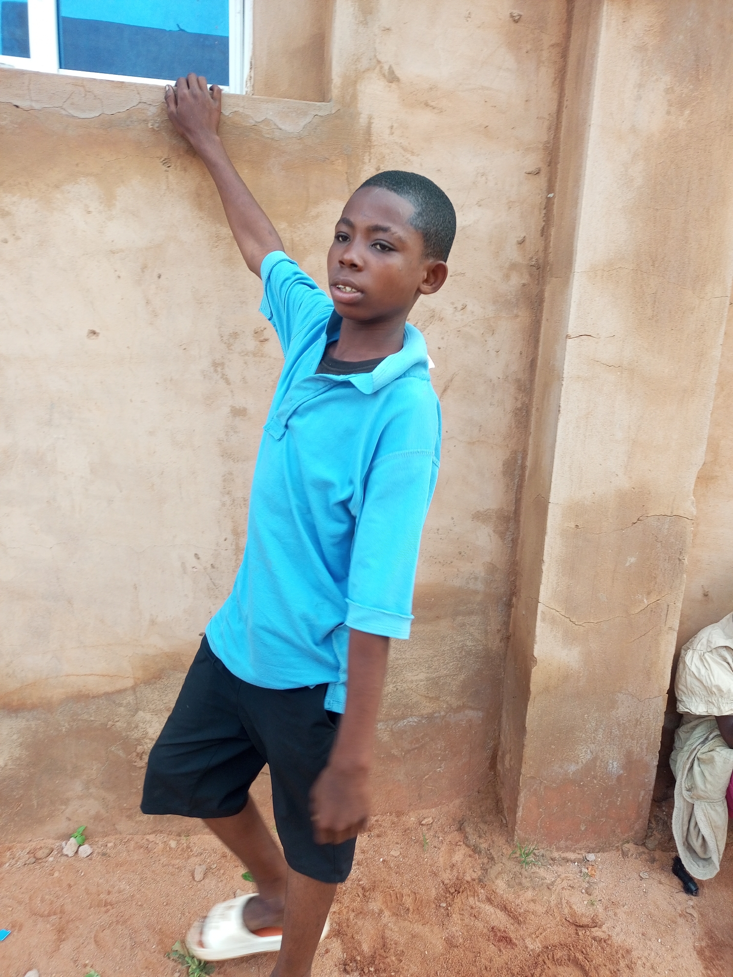 A young boy with dark skin and short hair stands outdoors, leaning against a textured, tan-colored wall. He is wearing a bright blue collared shirt and black shorts. His left arm is extended upwards, with his hand resting on the wall near a window with a blue pane. The ground is reddish-brown dirt with scattered small stones and a few blades of grass. To the right, another person is partially visible, seated and wearing a light-colored garment. The scene appears to be in an outdoor, possibly rural, setting. The image was taken on September 11, 2023, in Tempure, Nigeria.