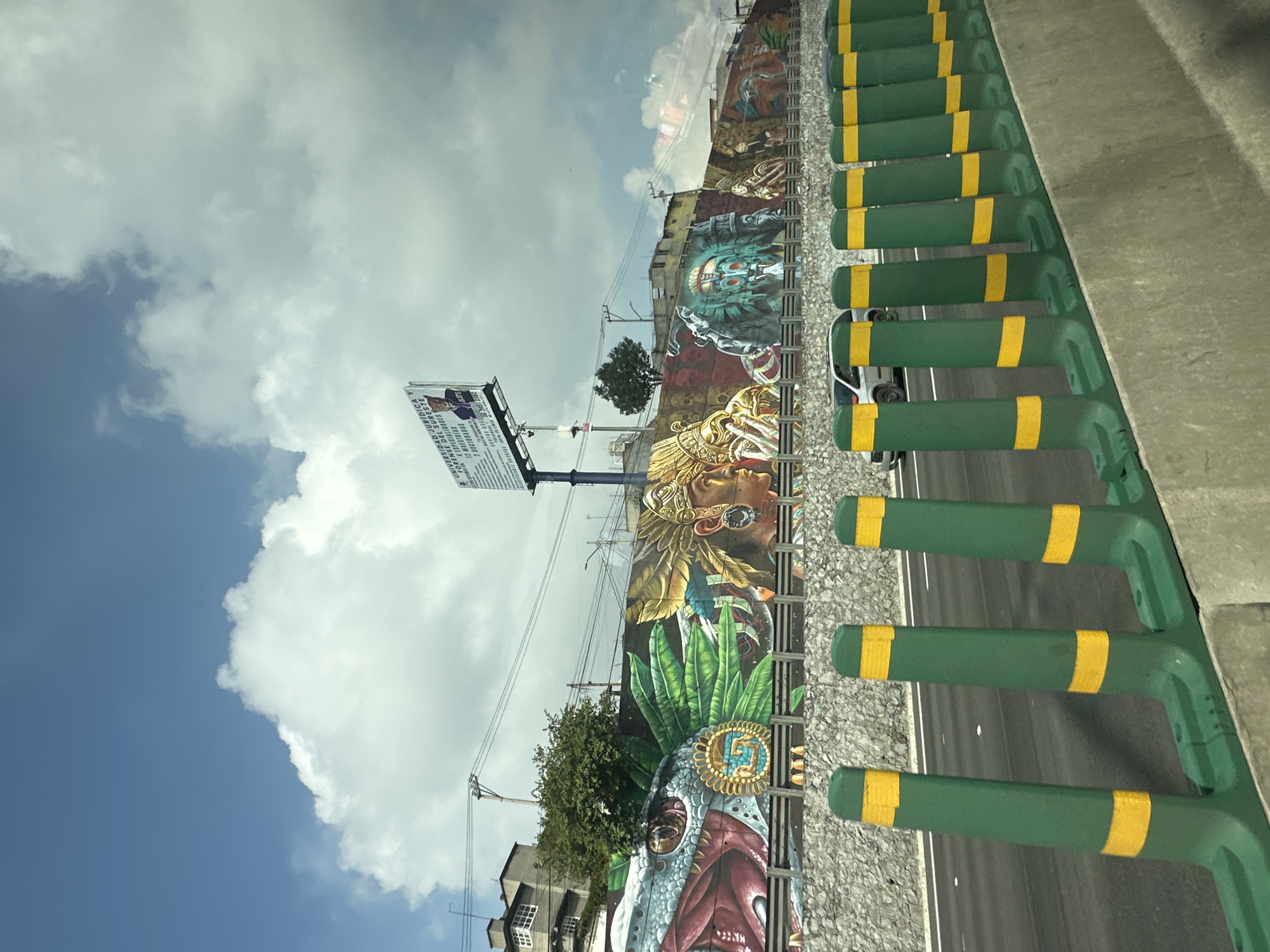 The image depicts a vibrant mural painted on a long, elevated wall alongside a highway in Valle de Chalco Solidaridad, Mexico. The mural features Aztec-inspired imagery, including a large serpent's head with intricate scales and a coiled headdress, followed by depictions of figures in feathered headdresses and ornate jewelry, with stylized skulls and masks interspersed. The artwork is colorful and detailed, showcasing a rich cultural heritage.
The highway itself is visible in the foreground, with a concrete barrier separating it from the mural. Green traffic delineators with yellow reflective stripes line the edge of the highway, indicating a traffic safety measure. A silver sedan is seen driving on the highway, partially obscured by the delineators.
Behind the mural, on the elevated terrain, there are buildings with windows and some trees. Power lines and poles are also present, suggesting an urban or semi-urban setting. The sky above is a bright blue with scattered white clouds. A large billboard for "UNIVERSIDAD JURIDICA FORENSE DEL SURESTE" is positioned above the mural, further reinforcing the urban context and advertising educational services. The overall scene captures a blend of historical art, modern infrastructure, and everyday life in the specified Mexican municipality.