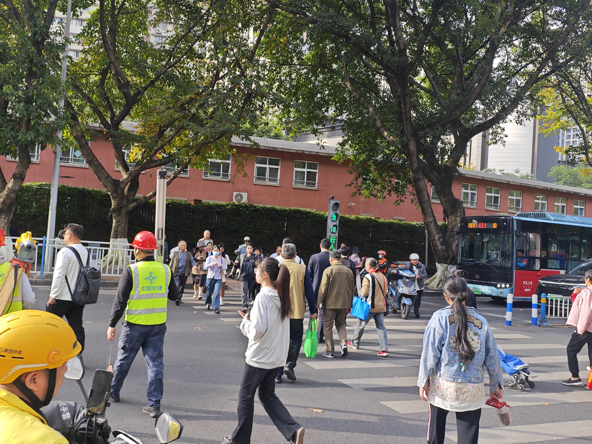 The image depicts a bustling street scene in an urban area, likely Yuzhong District, China, focusing on a crosswalk populated by numerous pedestrians. The time of day appears to be daytime, with clear weather and bright, natural lighting.
In the foreground, a person wearing a yellow helmet with "美团外卖" (Meituan Waimai, a food delivery service) is seated on a scooter or motorcycle, facing away from the camera. Beside them, on the crosswalk, stands a person in a bright green high-visibility vest and a red hard hat, with "中国交建" (China Communications Construction) written on the back of the vest. This individual seems to be overseeing or guiding pedestrians. Other pedestrians of various ages are actively crossing the street, some looking at their phones or carrying bags. Several people are wearing face masks. To the far left, partial view of surveying or construction equipment on a tripod is visible.
On the right side of the road, a blue public bus is visible, with the route number "421" clearly displayed, along with "重庆公交" (Chongqing Public Transport) and the bus number "84171". Another scooter is parked or moving near the bus. A green traffic light, indicating "行" (Go/Walk), is visible above the crosswalk.
The street is lined with mature trees featuring vibrant green foliage, partially obscuring the background. Behind the trees, a reddish-brown building with multiple windows can be seen, and taller, more modern white and grey buildings rise further in the distance. The overall impression is one of daily urban activity.