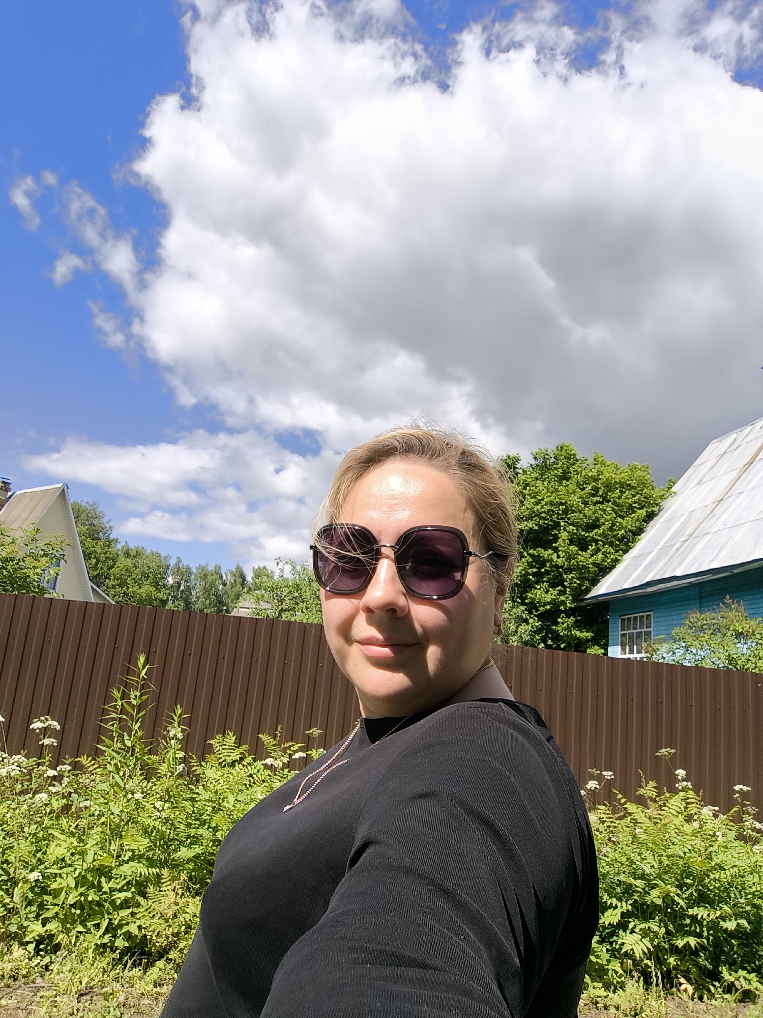 In a medium shot, a woman with blonde hair and sunglasses is seen in an outdoor setting. She is wearing a black top with a necklace. Behind her, a brown corrugated metal fence runs horizontally across the frame. Lush green foliage with small white flowers grows in front of the fence. In the background, trees, a house with a white roof and blue siding, and another house with a light-colored roof are visible. The sky above is blue with large, white cumulus clouds. The scene appears to be in the village of Novonikolskoye, Russia, likely during the daytime.