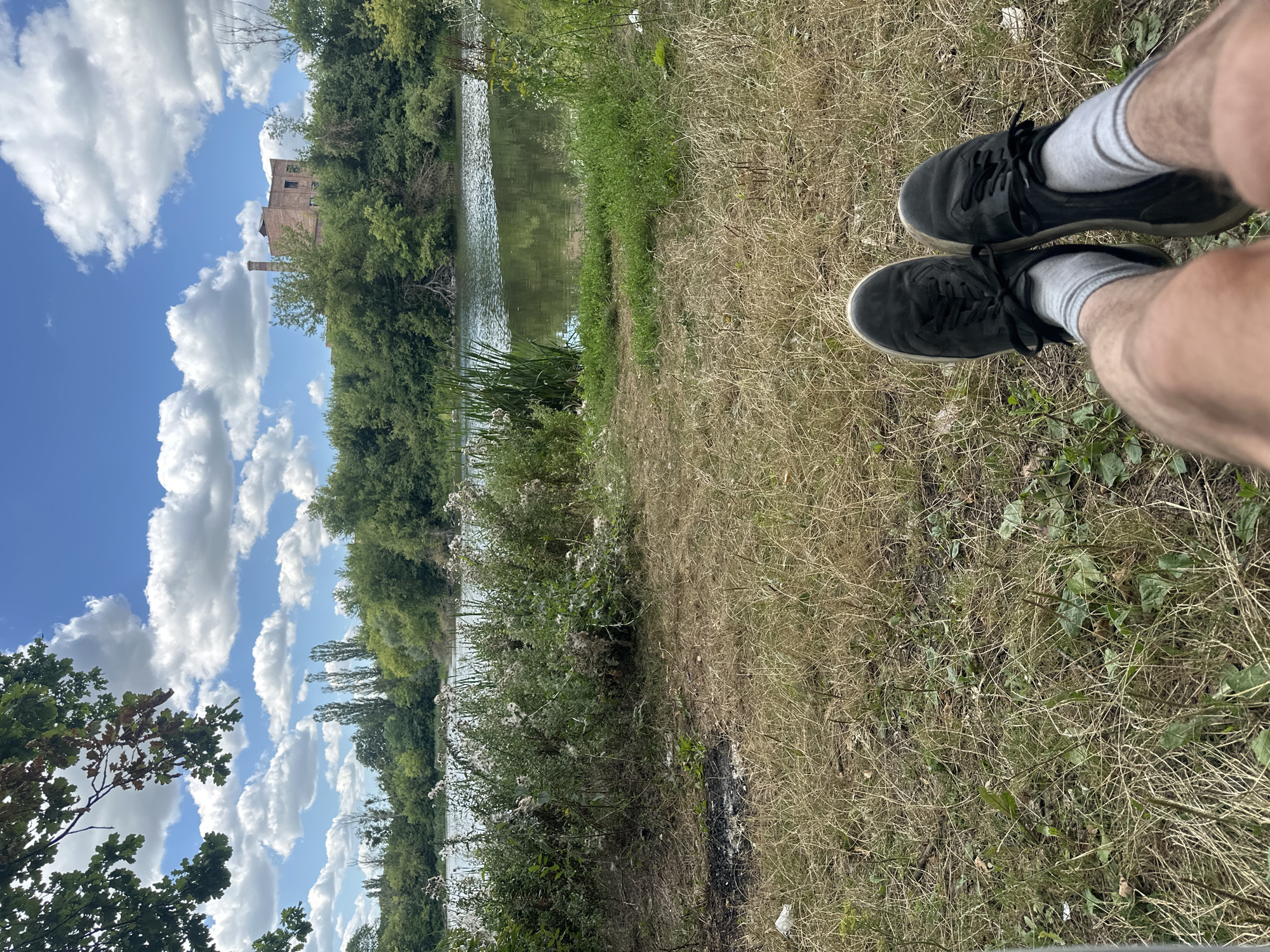 The image shows a body of water with trees and an old brick building on the far bank. In the foreground, a person's legs and feet wearing black sneakers and gray socks are visible, resting on dry grass. The sky is blue with scattered white clouds. The location is identified as Makiv, Ukraine.