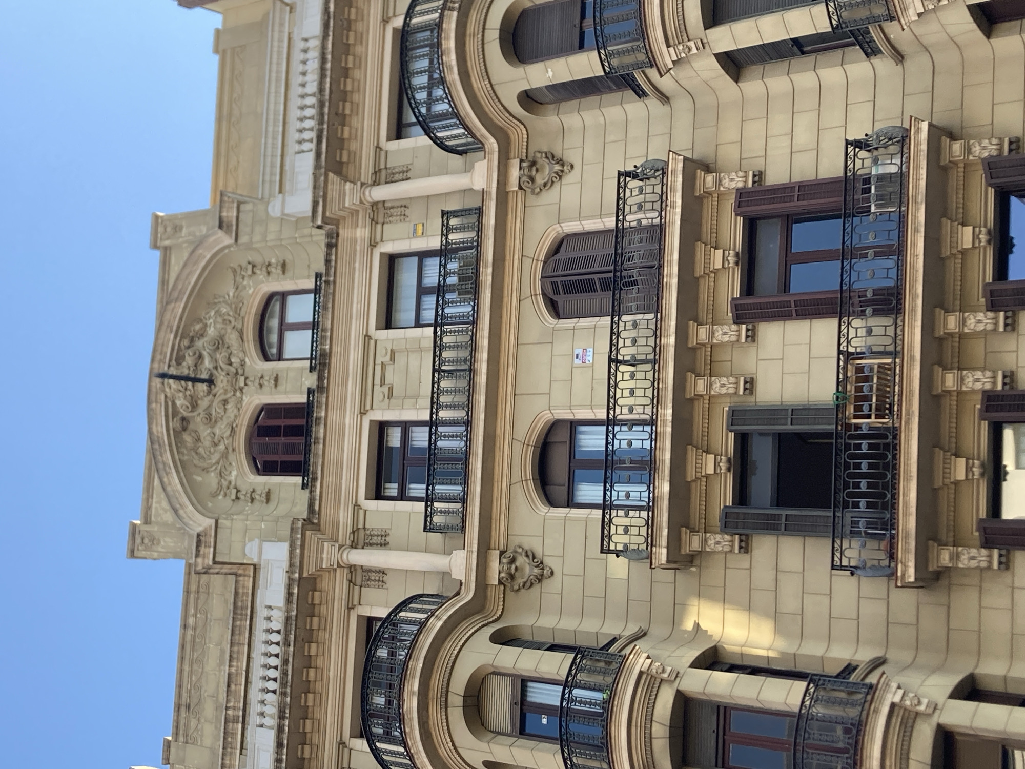 The image is a low-angle, upward-facing shot of an ornate building facade against a clear blue sky. The architecture appears to be in Barcelona, Spain, characterized by its elaborate detailing and neoclassical or Art Nouveau style. The building has multiple floors with numerous windows, many featuring dark-colored shutters and decorative wrought-iron balconies. Some balconies are curved, adding to the building's architectural flair.
The facade itself is a light beige or cream color with intricate carvings and moldings, including what appear to be lion or gargoyle-like decorative elements above some windows. The stonework is textured, with a grid pattern visible on large sections of the wall. The windows are mostly rectangular, but some are arched. The overall impression is one of grandeur and historical significance.
There is no visible text in the image. The time of day appears to be daytime, likely morning or afternoon, given the bright, even lighting and the clear sky, suggesting fair weather. The scene is a street view of a building, with no people or other objects visible that would indicate a specific activity or event. The focus is solely on the architectural details of the building.