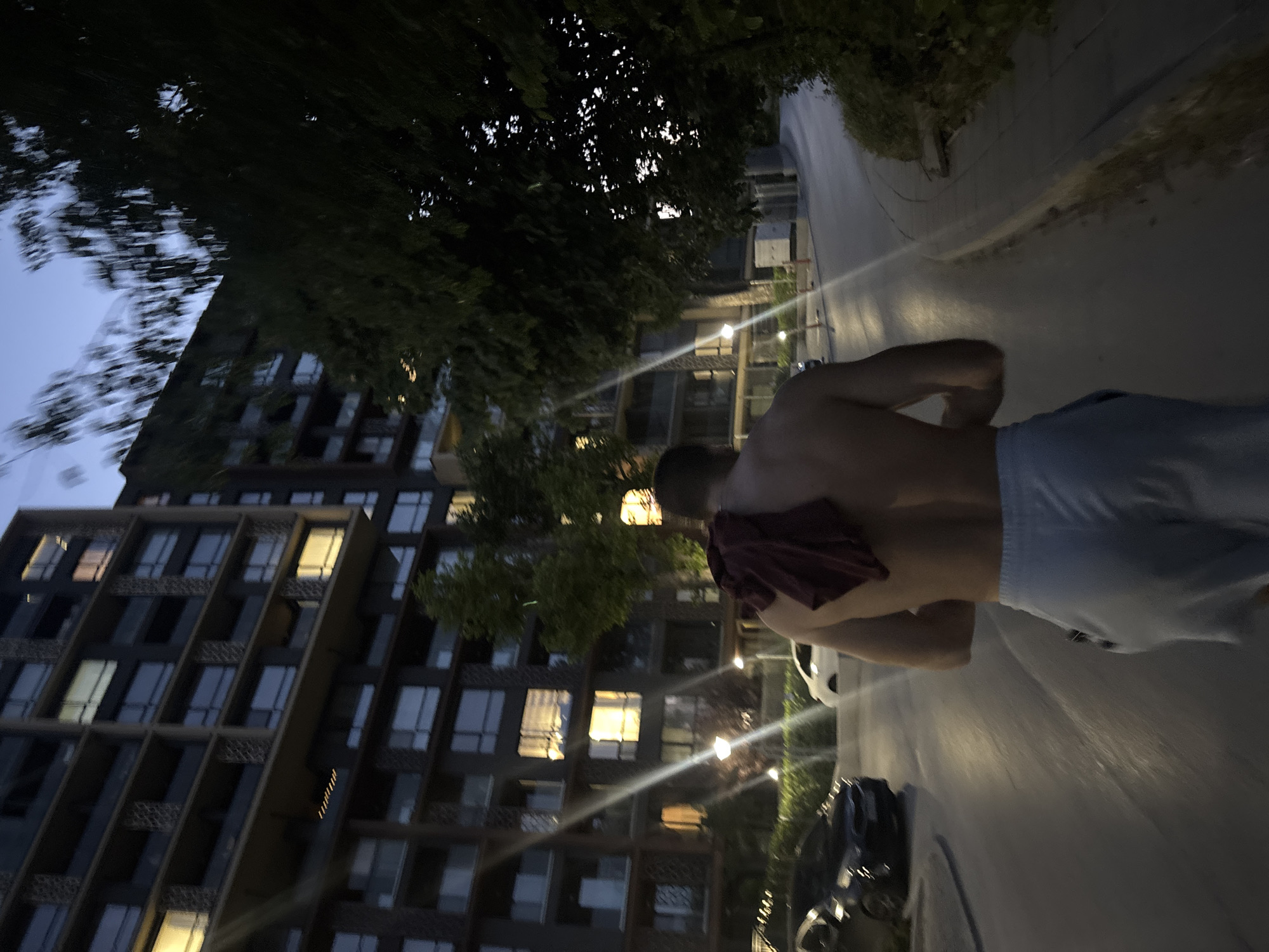 The image depicts a shirtless man with a dark red cloth draped over his right shoulder, walking away from the camera on a paved pathway. He is wearing light blue athletic shorts. The setting appears to be an urban environment at dusk or early evening, with a modern, multi-story apartment building with illuminated windows on the left side of the frame. Trees with dense foliage are visible on the right side of the pathway and behind the building. The pathway is lined with streetlights, and a few cars are parked or driving on the road visible to the left of the pathway. The overall atmosphere suggests a residential area within the city of Varna, Bulgaria.