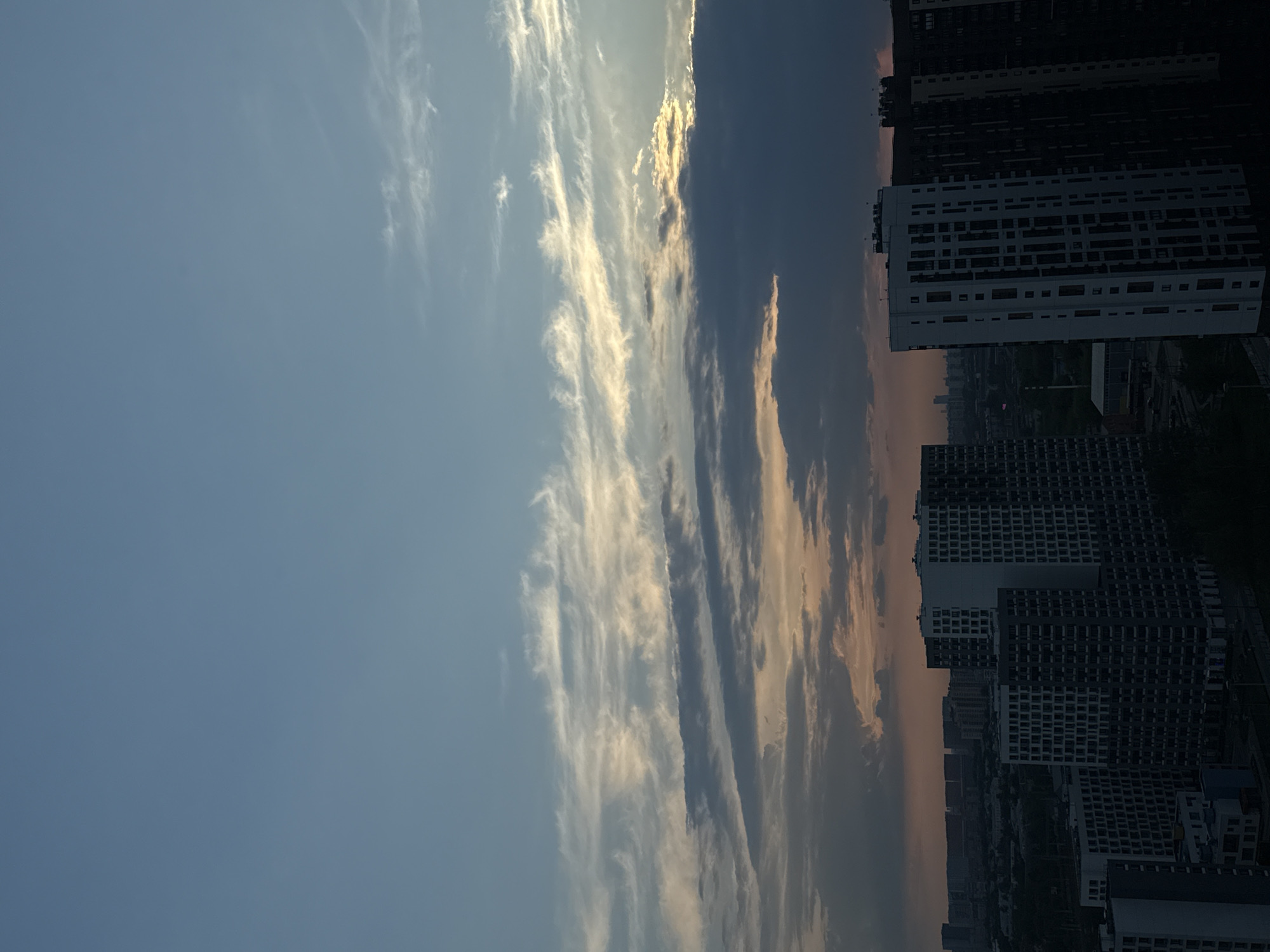 The image shows an urban landscape in Lyubertsy, Russia at dusk. The view is vertically oriented. The sky transitions from a clear blue to a darker, cloudy area with streaks of illuminated clouds. Below the sky, there are several tall apartment buildings of varying heights and designs. The buildings are densely packed, indicating a high population density. The overall ambiance suggests a late evening or early morning. The image lacks any identifiable people or specific activities.