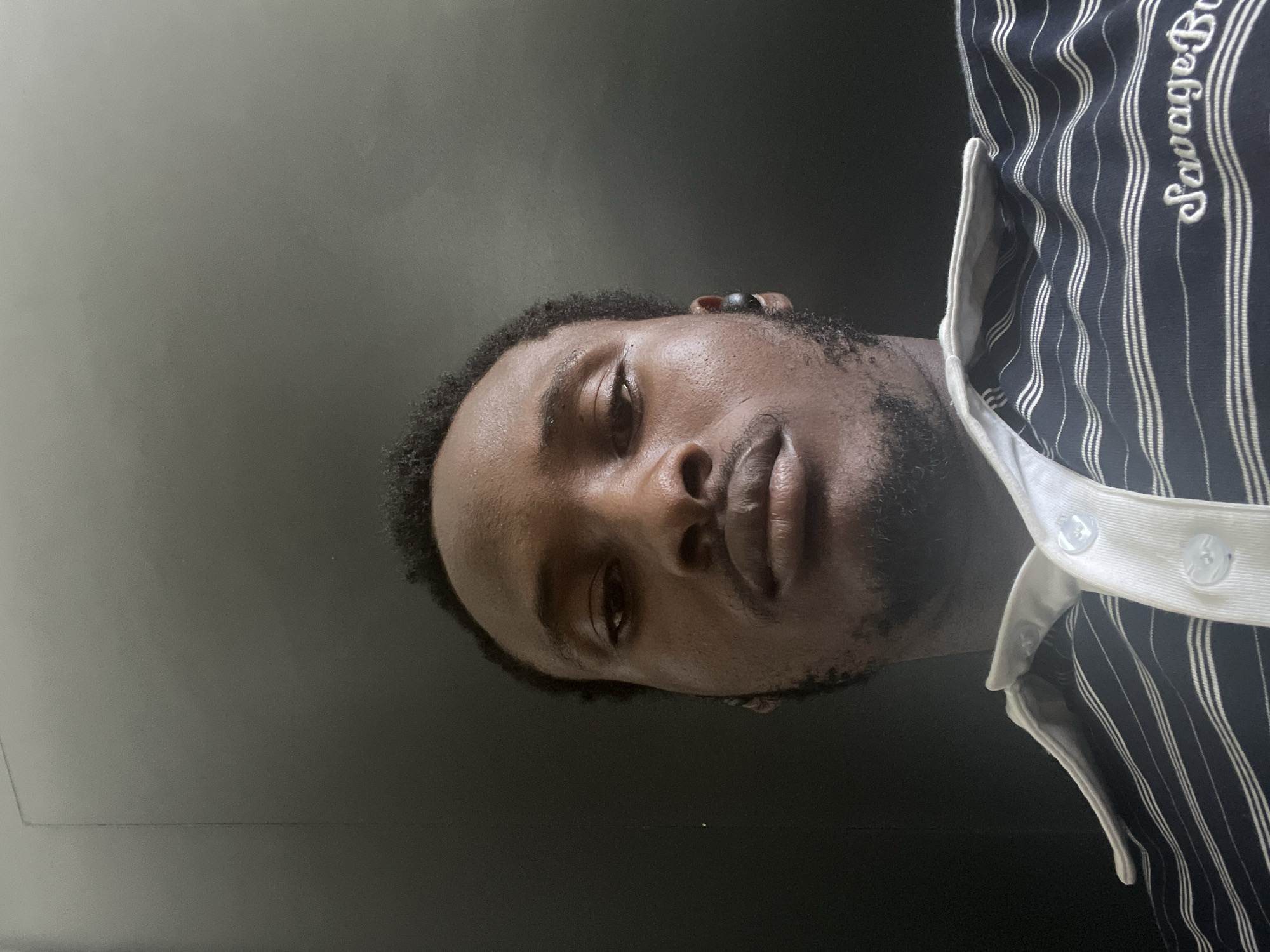 A medium shot depicts a person with dark skin and short, dark, curly hair, likely a man, facing the camera at a downward angle. He has a short beard. The subject is wearing a navy blue striped shirt with white pinstripes and a white collar. The shirt has the words "Savage Br" embroidered in white on the upper right. The background appears to be a dark grey or black surface. There is no explicit evidence of the location being Lugbe, Nigeria. The details of the image do not provide enough information to establish the location.