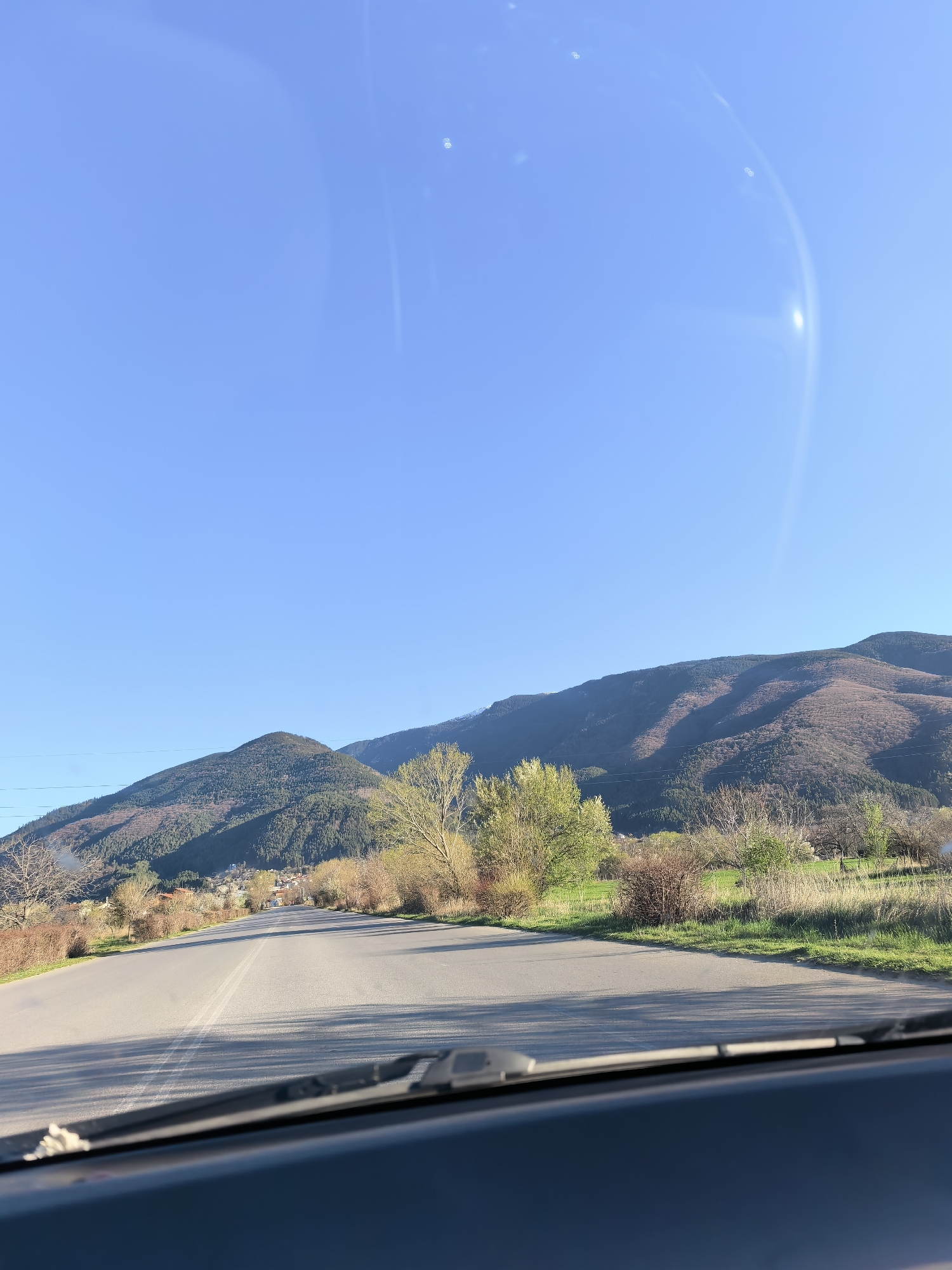The image depicts a view from inside a vehicle, looking out onto a road leading toward a mountain range under a clear blue sky. The road is grey asphalt with white lines marking the lanes. Vegetation, including trees and shrubs, lines both sides of the road, some displaying green foliage. The mountains in the background are covered in vegetation, with variations in color suggesting different types of trees or stages of growth. Overhead power lines are visible to the left of the frame. The bottom of the image shows the dashboard and windshield wipers of the vehicle. The location is unknown.