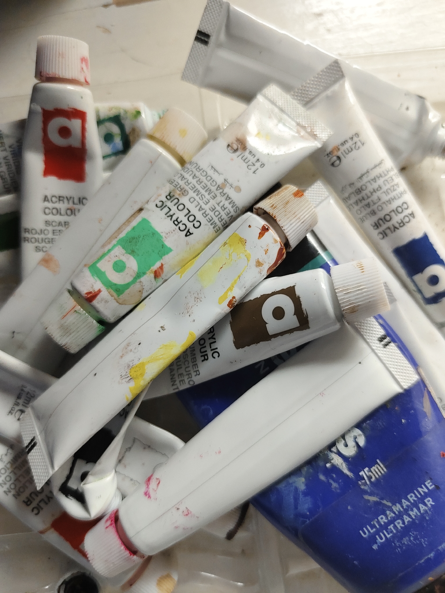 The image displays a cluttered assortment of approximately ten to twelve used acrylic paint tubes, predominantly white with various colored labels and caps, piled haphazardly on a light-colored, possibly off-white, textured surface. No discernible elements in the image indicate its location in Davao City, Philippines.
The subjects are mostly small tubes, many marked "12ml e / 0.4 US fl.oz", and one larger blue tube marked "75ml" with "ULTRAMARINE" and "ULTRAMAR" printed on it. All visible tubes bear the brand "ACRYLIC COLOUR" and feature a stylized lowercase 'a' logo within a distinct colored square corresponding to the paint hue. Specific colors identifiable include a red tube ("ROUGE SCARLET RED" partially visible), a green tube ("EMERALD GREEN" partially visible), a blue tube ("PHTHALO BLUE / BLEU PHTHALOBLAU" partially visible), and a brown tube (partially visible, likely "UMBER"). Another tube shows a black logo.
The tubes exhibit signs of use, including squeezing, creasing, and smudges of dried paint on their bodies and caps. Some caps are white, others are stained with their respective paint colors, such as red on a white cap, and yellowish-brown on another. Text is visible in English, French, Spanish, and Arabic on various tubes. The tubes are piled in different orientations, suggesting they have been recently used and placed down. The background beyond the immediate pile is indistinct and light-toned. No individuals are present in the scene.