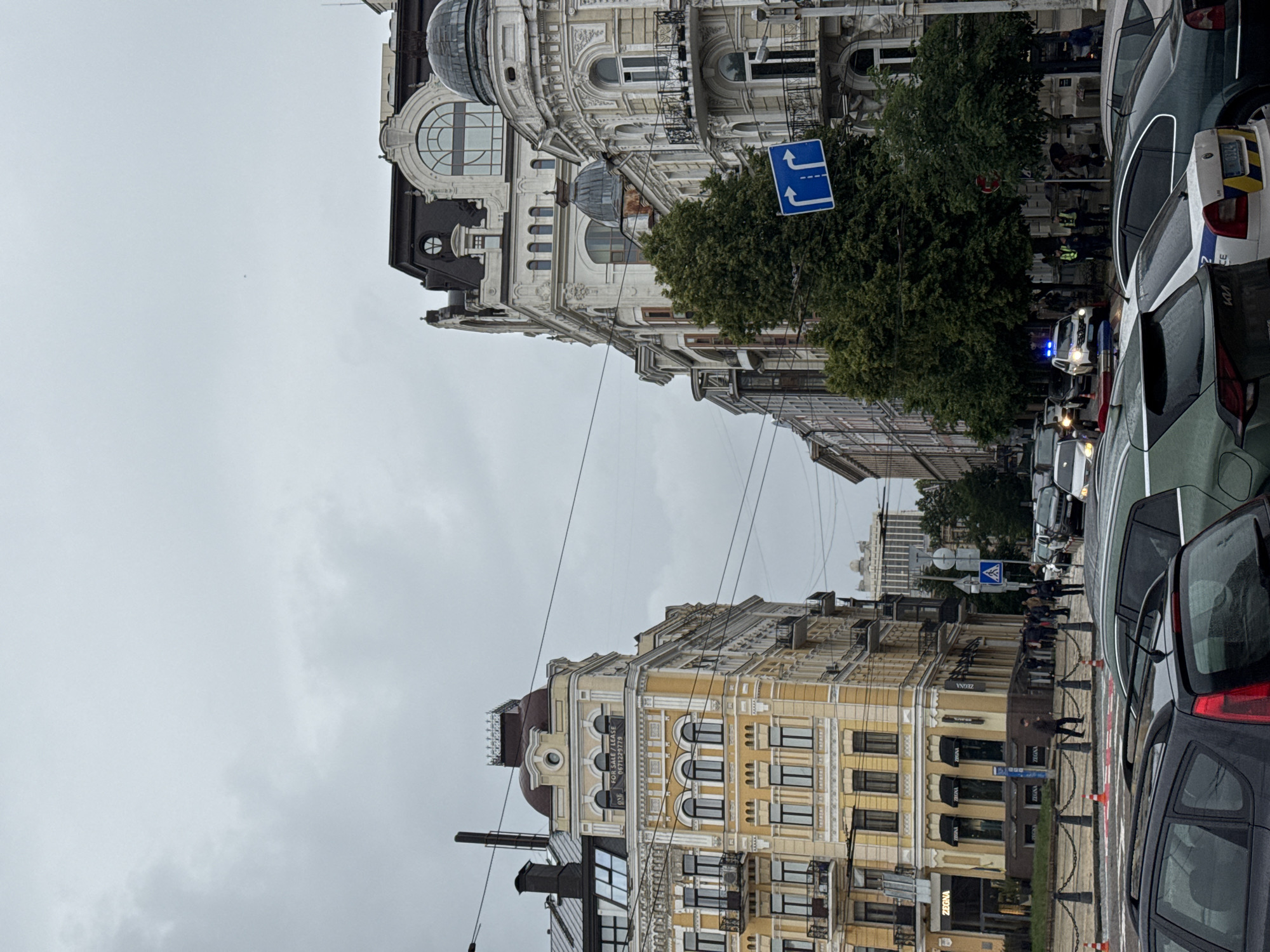 The image captures a vibrant city street in Kyiv, Ukraine, under an overcast sky, suggesting daytime. The street is flanked by ornate, multi-story buildings exhibiting classical architecture. On the left, a grand light-colored building stands out with its intricate decorative stucco work, large arched windows, balconies, and prominent domes. On the right, a similar historical building, predominantly yellow, also features detailed facades and arched windows.
The street itself is busy with numerous vehicles, including cars parked along the sidewalks and others seemingly in traffic. A notable event unfolds in the mid-ground, where a police presence is evident. A police vehicle with flashing blue lights is visible, alongside a white car with the word "POLICE" partially legible on its rear. Several uniformed individuals are seen near these vehicles, gathered with a small group of people, indicating a potential incident or official activity.
Pedestrians are visible on the sidewalks, moving about their day. Green trees are planted along the street, adding foliage to the urban landscape. Overhead utility wires crisscross the view. Street furniture includes orange and white traffic cones and a blue directional road sign displaying two upward arrows, indicating lanes for straight travel.
Visible text on a banner attached to the yellow building reads "FOR SALE / LEASE 09711229779." Additionally, store signage below the yellow building displays the brand "ZEGNA." The scene offers a glimpse into daily life in a historic European city, marked by its distinctive architecture and urban activity.