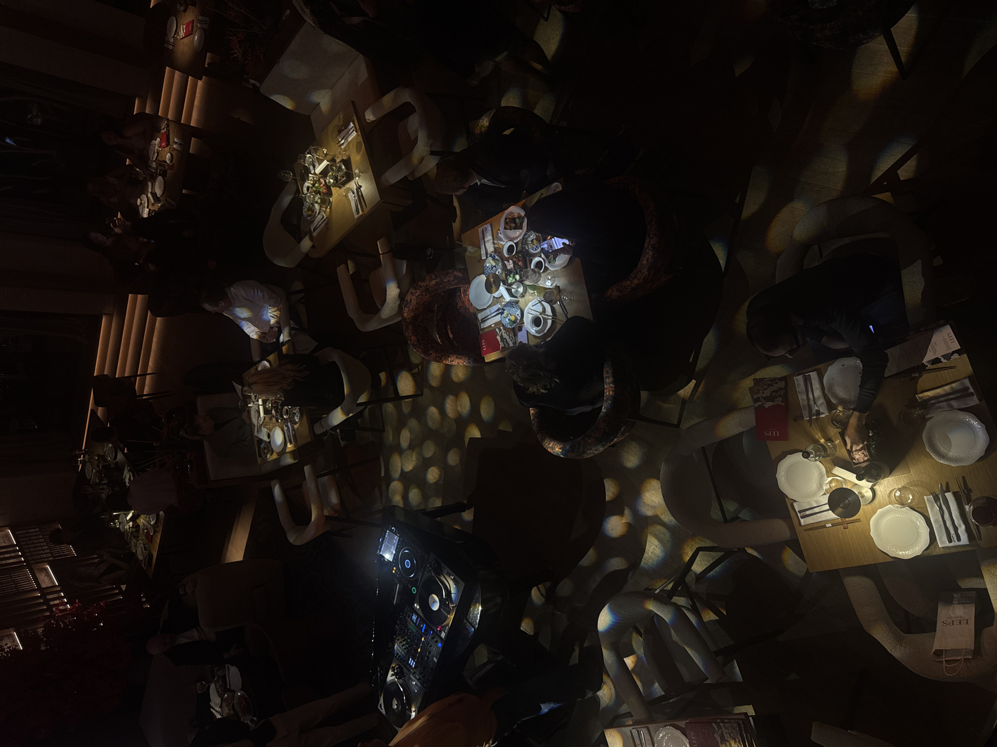 This is a high-angle, overhead shot of a dimly lit, upscale restaurant or lounge, likely taken during the evening or night. The scene is dominated by deep shadows, with focused spotlights illuminating various wooden tables and their occupants. Decorative circular light patterns are projected onto the floor and surrounding surfaces, creating a dynamic ambiance.
Several individuals are seated at tables set with white plates, cutlery, and an assortment of glassware for drinks. Some tables feature small bowls of food, possibly appetizers or sushi. Guests are seen interacting, and some are looking at their mobile phones, whose screens cast a soft glow.
A prominent professional DJ mixing console, illuminated with various lights and featuring turntables, is visible in the foreground, suggesting the presence of live music or a club atmosphere. The chairs around the tables vary in design, including upholstered and more minimalist styles.
The word "LEPS" is visible on several menus or booklets placed on the tables, and also on a paper bag, likely indicating the name of the establishment. The overall atmosphere is sophisticated and social, typical of a vibrant nightlife or dining venue in Moscow, Russia.