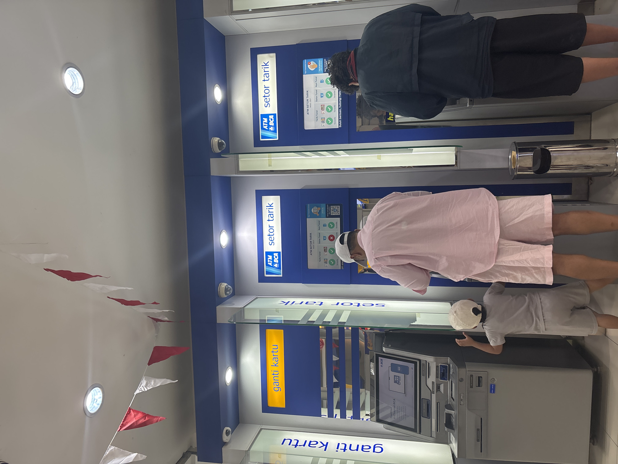 In North Jakarta, Indonesia, at an ATM bank located inside a building with fluorescent lighting, three individuals are observed. One person, wearing a light pink polo shirt and matching shorts, stands in front of an ATM machine. To their left, a young child in a grey t-shirt and shorts faces the same ATM. To the right of the person in pink, another individual, dressed in a dark blue jacket and black shorts, is using a different ATM machine. Red and white triangular flags are strung across the ceiling, possibly for a national celebration. The ATMs have signage indicating services like "ganti kartu" (change card) and "setor tarik" (deposit/withdraw).