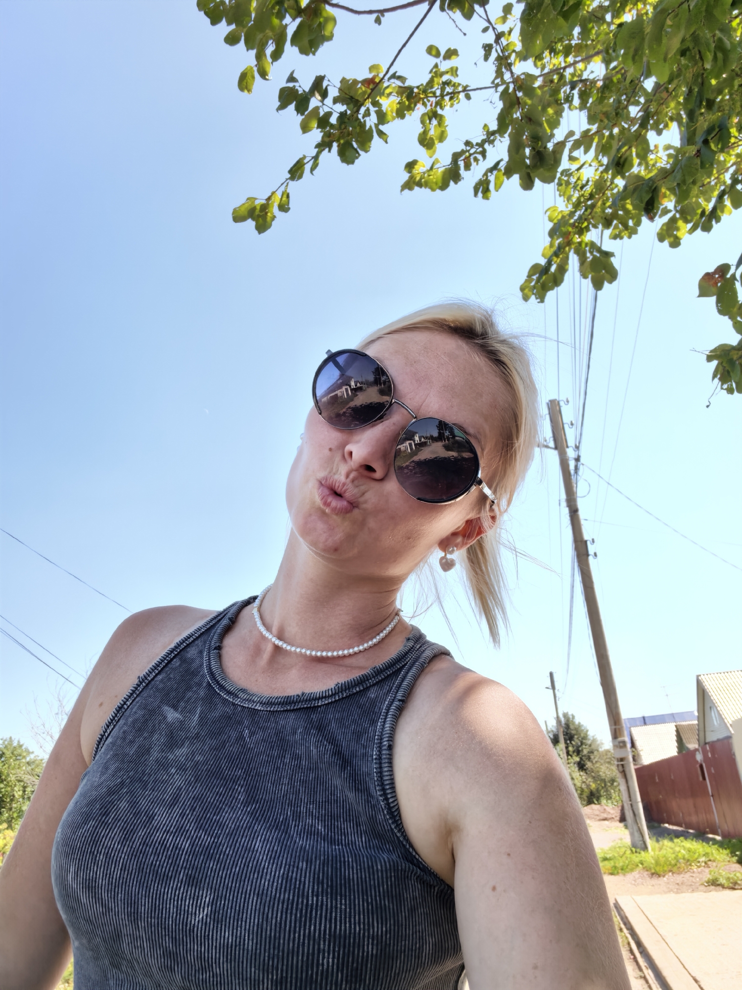 A blonde woman wearing sunglasses and a pearl necklace takes a selfie outdoors. She is wearing a grey ribbed tank top and has her lips pursed. In the background, there are trees, a utility pole, and houses. The sky is blue and clear. The scene appears to be in a rural or suburban area. There is no indication that the location is Sterlitamak, Russia.
