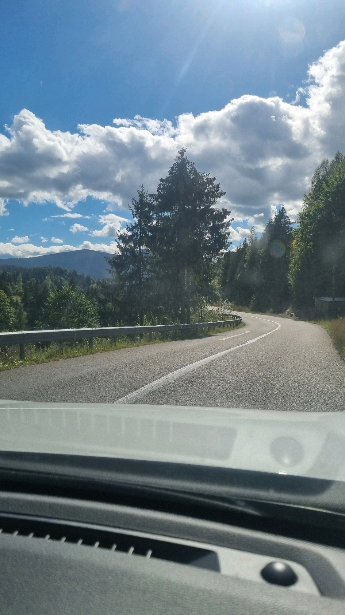This is a first-person perspective image taken from inside a vehicle, looking out at a winding road through a forest with mountains in the distance under a partly cloudy blue sky. The road has a white line marking its center and right edge, and a metal guardrail runs along the left side. The foreground shows the interior of the car, including part of the dashboard. The scene is set in a natural, mountainous environment. There are no people visible in the image. The weather appears to be sunny with some scattered clouds.