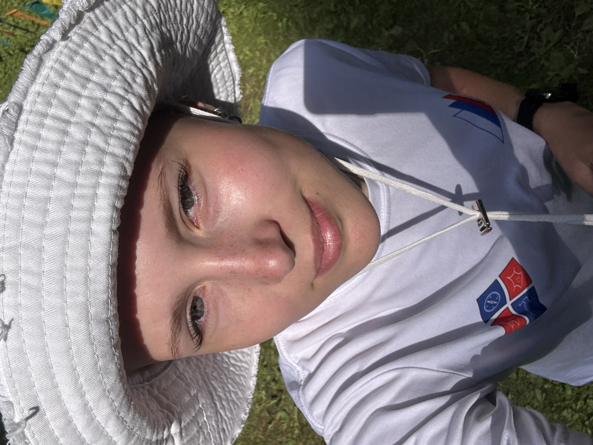 This is a close-up, rotated portrait of a young person wearing a white bucket hat and a white t-shirt. The person is looking towards the camera with a soft, natural expression. Their eyes are light-colored, and they have light brown eyelashes and eyebrows. A few freckles are visible on their nose and cheeks. The person is wearing a white t-shirt with a blue and red emblem on the chest, possibly a logo or crest. A white drawstring is visible around their neck. The background consists of green grass, suggesting an outdoor setting. The lighting indicates it is daytime, likely sunny, casting a shadow from the hat onto the person's face. The overall impression is one of a casual, relaxed moment outdoors.