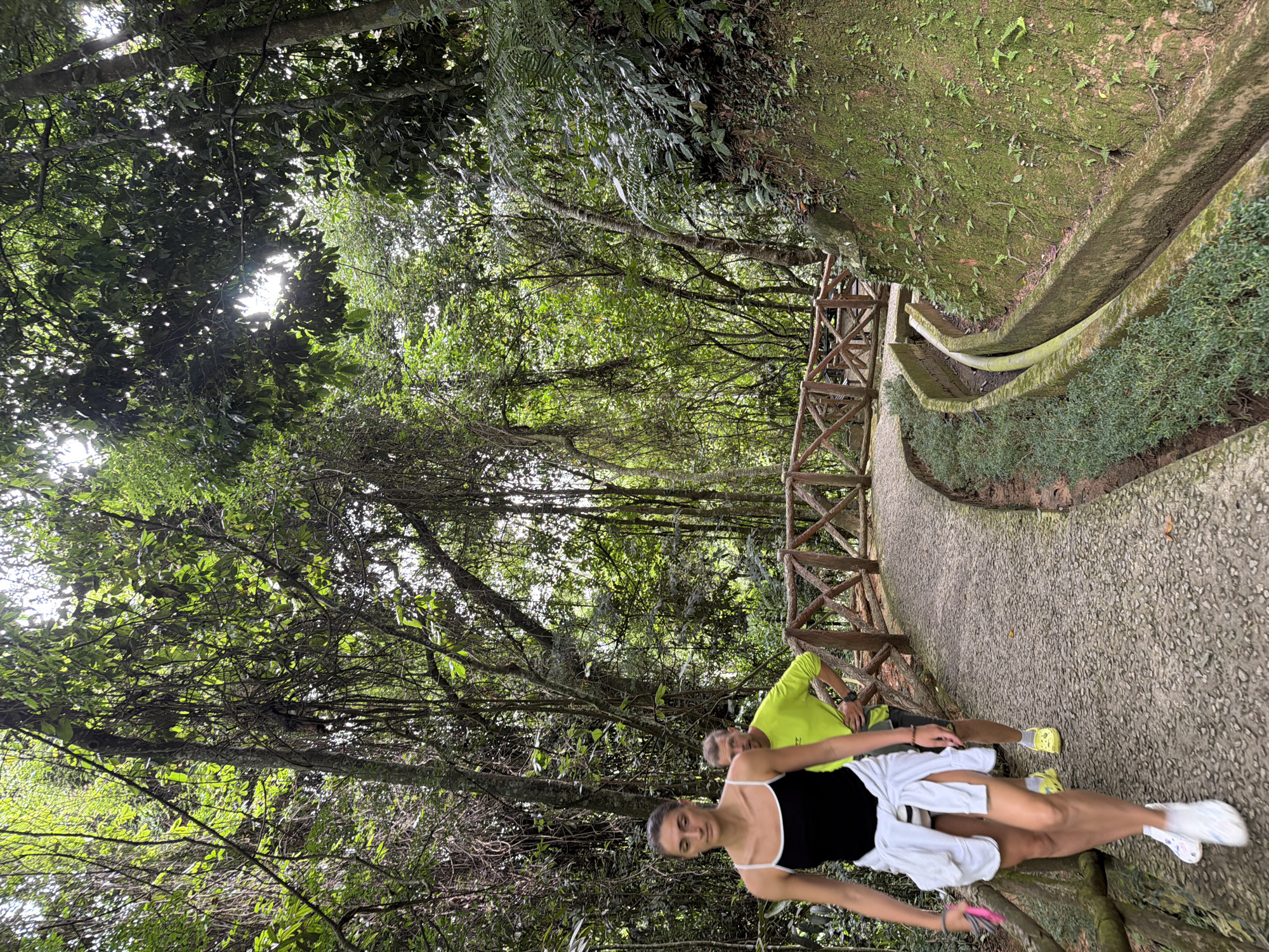 Two individuals are walking on a paved path in a lush, green forest. The woman in the foreground is wearing a black tank top, white shorts, and white sneakers, with a white jacket tied around her waist. She is looking forward and to her left. Behind her, a man in a bright yellow long-sleeved shirt and dark shorts is walking, looking towards the camera. The path curves to the right, bordered by a wooden railing. The environment is dense with trees, vines, and foliage, suggesting a natural park or botanical garden setting. The sun is filtering through the canopy, creating dappled light. The scene is consistent with a walking trail in the city of Da Lat, Vietnam, known for its cool climate and extensive pine forests and botanical gardens.