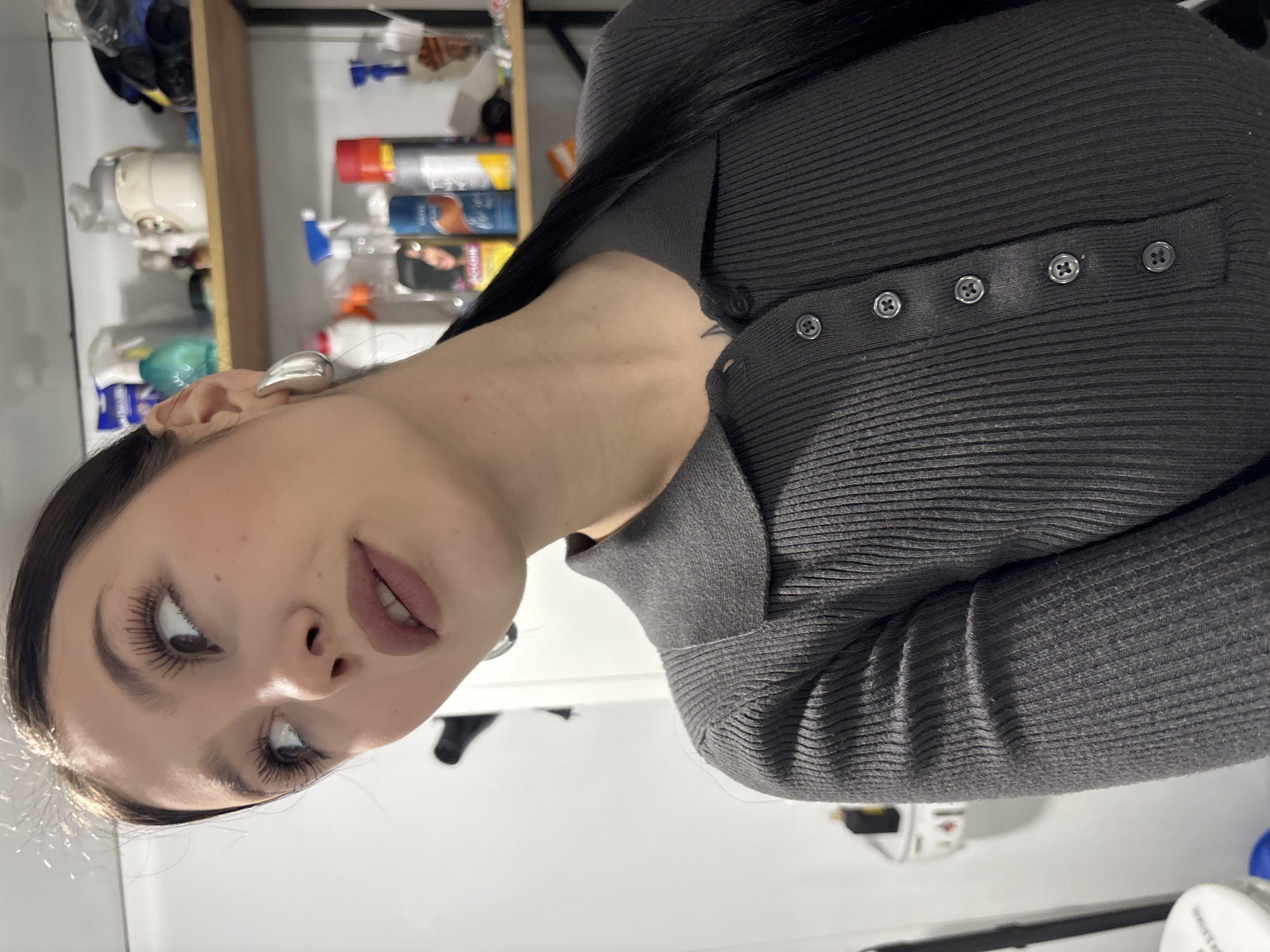 A woman is shown from a low angle. She has long, dark hair and is wearing a gray, ribbed top with buttons. The background includes a shelf with various household items and cleaning supplies, and a white surface. The location is not apparent from the image, therefore I cannot provide location data for Фоки, Russia.