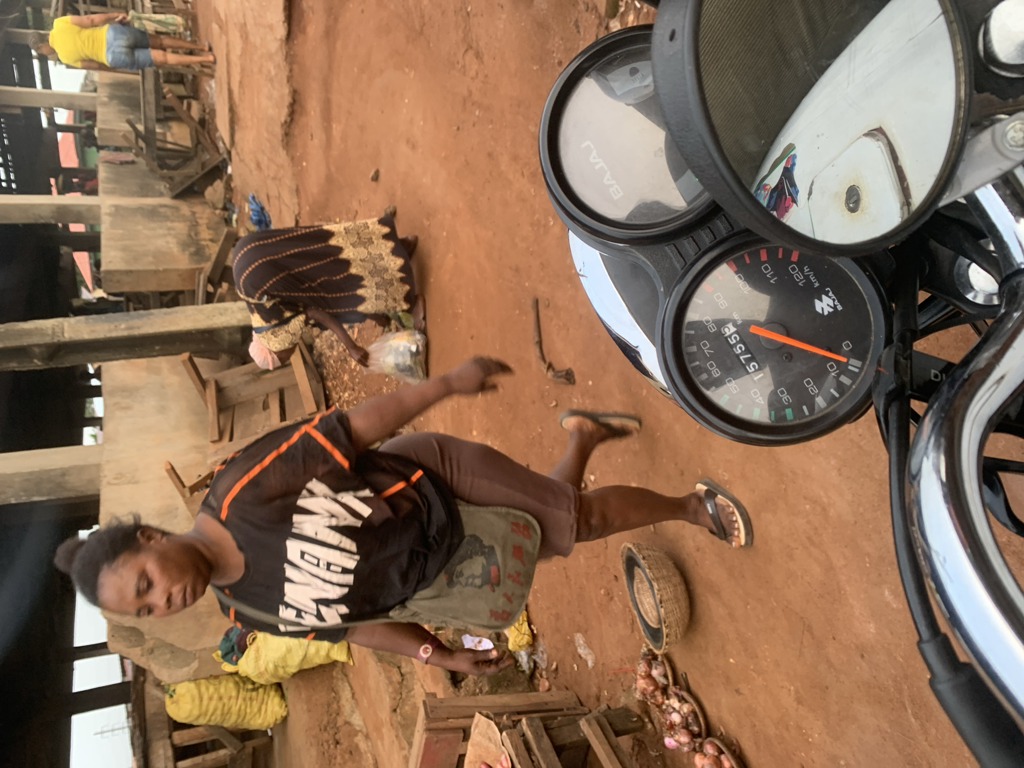 The image depicts a street scene in the city of Shagamu, Nigeria. In the foreground, a motorcycle's dashboard is visible, with a speedometer showing a reading of 15755 km/h and a BAJAJ logo. A woman in a black t-shirt with orange trim and brown shorts is walking in the middle ground, carrying a green messenger bag with a portrait on it. To her left, there are plates of onions and a woven basket. In the background, another woman in a dark dress with gold trim is bending down, holding a plastic bag. A woman in a yellow top and denim shorts is walking away in the distance. The environment appears to be an outdoor market, with wooden stalls and structures suggesting a commercial area. The ground is covered in reddish-brown dirt.
