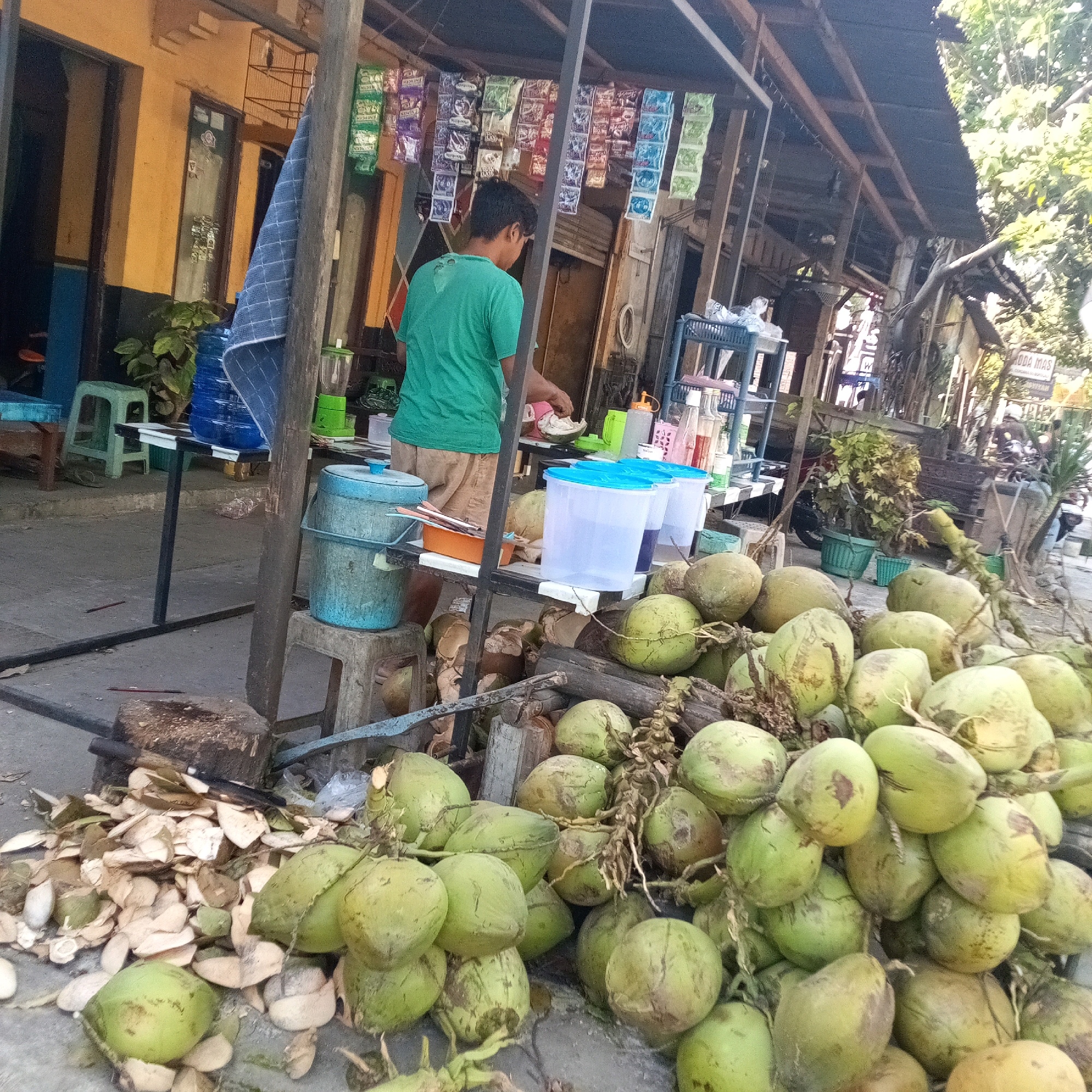 A young man in a green t-shirt and brown shorts is preparing coconuts at a street stall in Boyolali, Indonesia. He is standing behind a table with several large plastic containers of water and is in the process of husking a coconut. In front of the stall, a large pile of green coconuts and coconut husks are spread on the ground. Behind the young man, a variety of products are displayed on shelves, including bottled drinks and packaged snacks. The stall appears to be made of wood and metal and is covered by a dark-colored roof. The background shows a building with yellow walls and windows. The scene is illuminated by daylight.