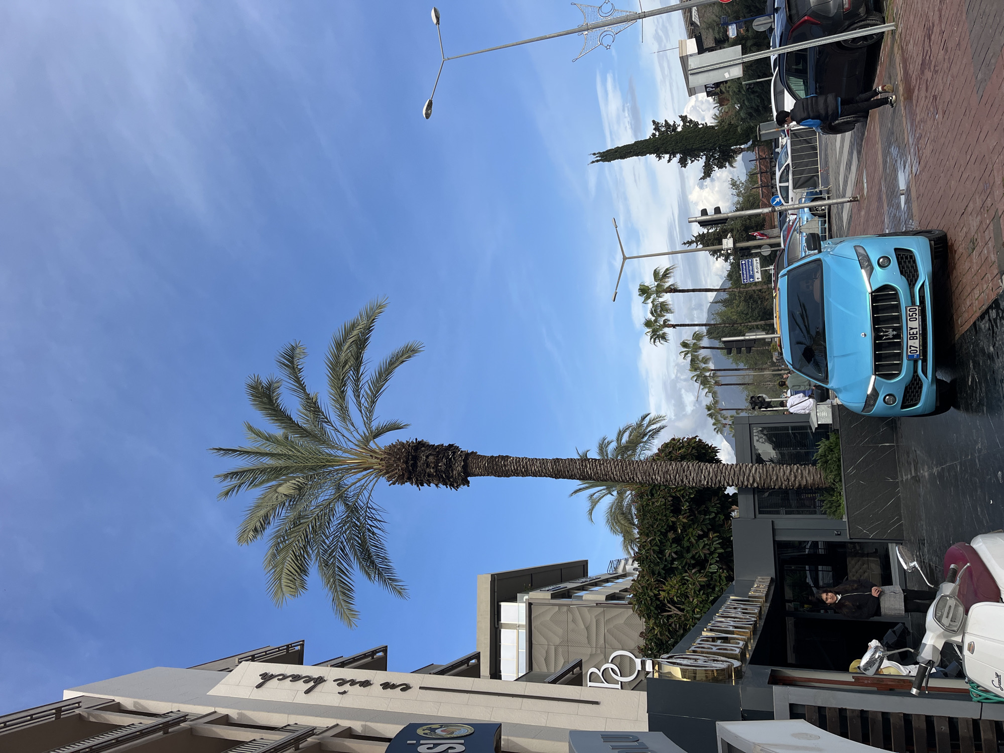 The image depicts a street scene in Alanya, Turkey, with a blue Maserati car prominently parked on the right side of the frame. The license plate reads "07 BEY 050." A person is standing near the car. A palm tree stretches into a bright blue sky. The architecture includes modern buildings with the text "Bohemian sea beach" visible on one. There are street lights, other palm trees, and distant hills in the background. A person is sitting in front of a building, likely the entrance to a business. Several scooters are parked on the right, near the car. The pavement appears wet, suggesting recent rain or cleaning.