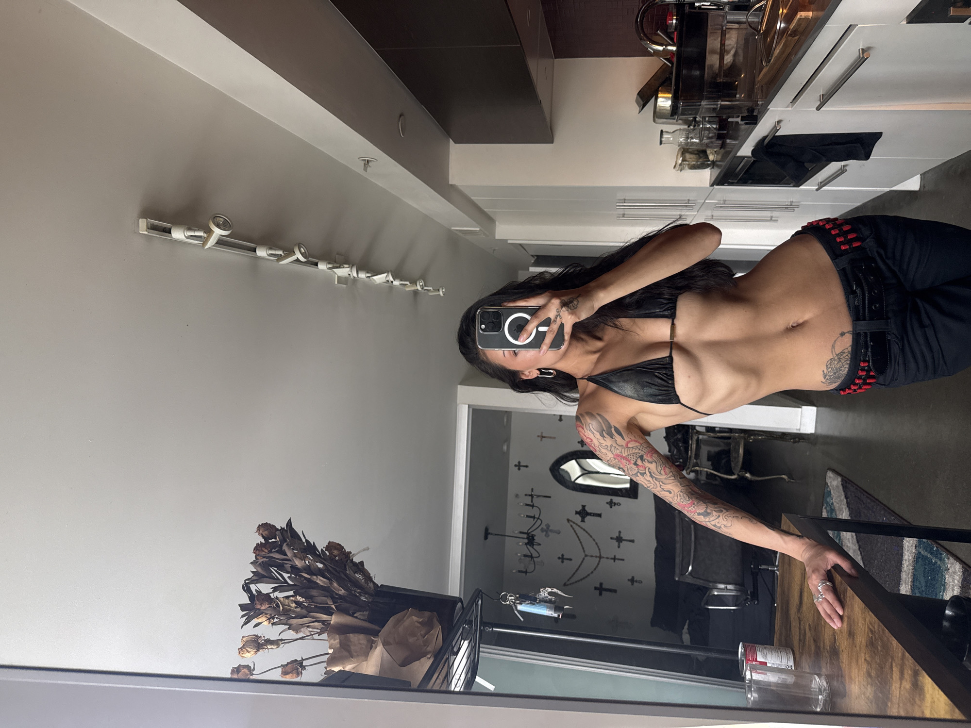 A selfie is taken in a mirror, featuring a person with long dark hair, wearing a black halter-neck top and black pants with a belt adorned with red, bullet-like embellishments. Extensive, colorful tattoos cover their left arm, showcasing dragon and floral designs, with additional black/grey tattoos on their abdomen and hip. They hold a smartphone in a clear MagSafe case, partially obscuring their face, while their left hand, decorated with rings, rests on a wooden table.
The interior setting, possibly a modern apartment or loft in Vancouver, Canada, is reflected. To the left, a light grey wall features contemporary track lighting, with a darker bulkhead above. The right side reveals a sleek kitchen equipped with white cabinets, silver handles, and stainless steel appliances. A black cloth is draped over a cabinet handle.
On the table in the foreground are dried flowers in a container, a clear glass, and a can partially visible with an "S" on its label. Beyond the kitchen, a doorway leads to an adjoining room, characterized by a dark wall decorated with numerous cross motifs, an arched mirror, and a chandelier. A patterned rug lies on the floor, completing the modern aesthetic with subtle gothic touches. The activity is the person capturing their reflection, likely in a full-length mirror.
