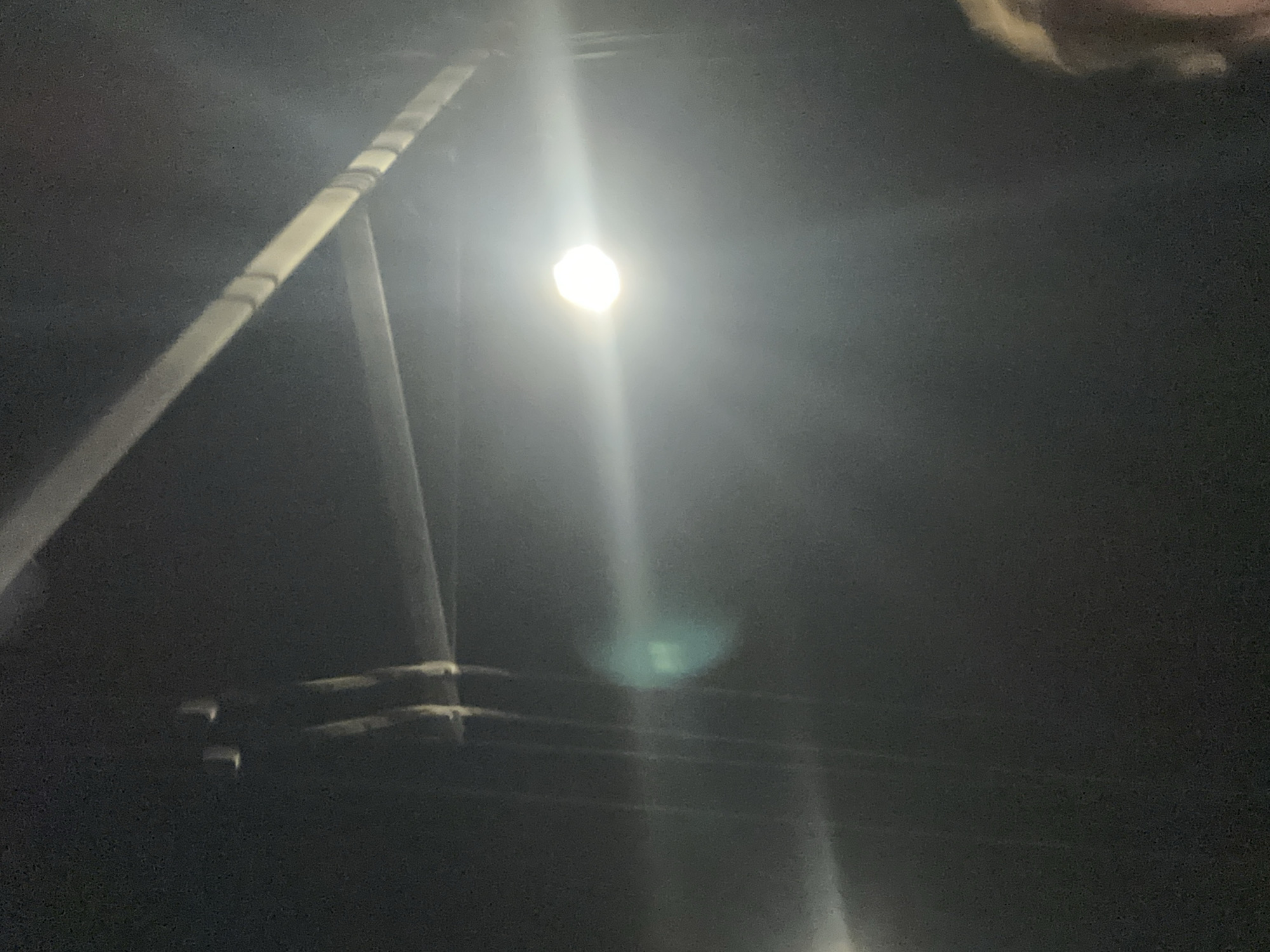 The image shows a nighttime view of a street scene with a bright street lamp illuminating the surroundings. A utility pole, with attached lines and hardware, is visible on the left side of the frame. The image is dark with limited details due to the low light conditions. The location is unknown.