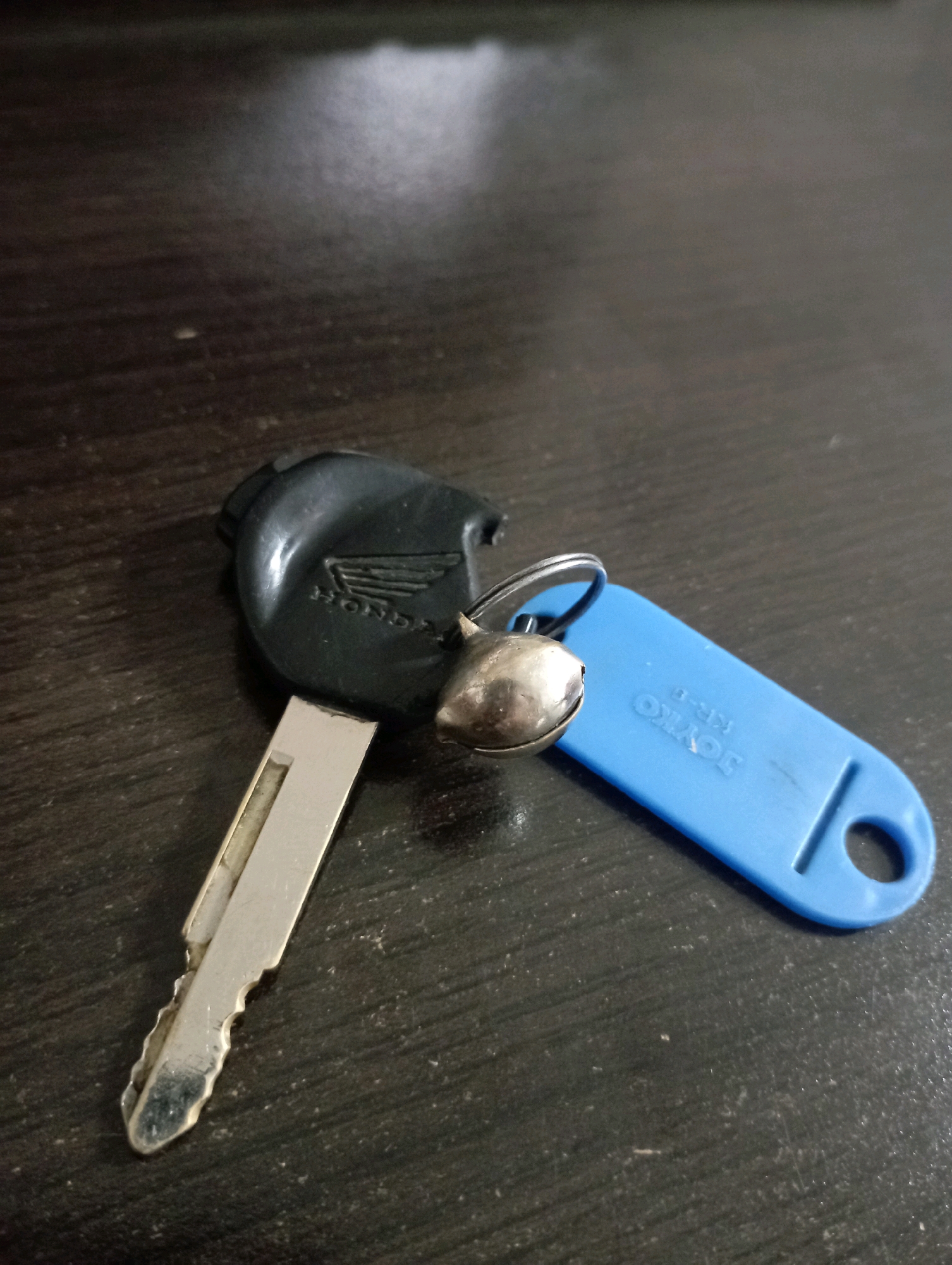 A Honda motorcycle key with a small silver bell and a blue plastic tag lies on a dark, wooden surface. The key has a black plastic head with the Honda logo embossed on it. The key's metal blade is silver and has visible cuts and notches. The blue tag is oval-shaped with a hole at one end and text that appears to read "JOYKO KR-B". The wooden surface has a visible grain pattern and some dust particles are present. The lighting suggests an indoor environment, possibly a table or desk. The location is not specified in the image itself, but based on the prompt, it is stated to be in Ponorogo, Indonesia. There are no people or interactions visible in the image.