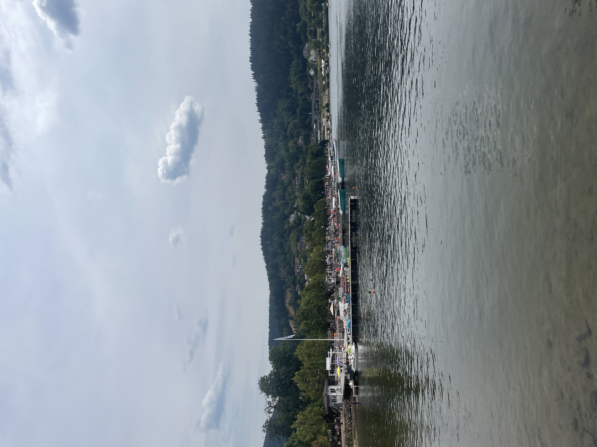 This image depicts a tranquil scene at the lake in Gérardmer, France. The foreground is dominated by the calm, clear water of the lake, with the sandy bottom visible beneath the surface in shallower areas. In the mid-ground, a wooden pier extends into the lake, lined with various boats, including small pedal boats and motorboats. Some people are visible on the pier and in the water, some of whom appear to be swimming. Beyond the pier, the shore is lined with trees and buildings, including a small kiosk-like structure. The background features a dense, tree-covered hillside dotted with houses, reaching up to a cloudy, overcast sky. The overall impression is one of a pleasant, relaxed day by the water.