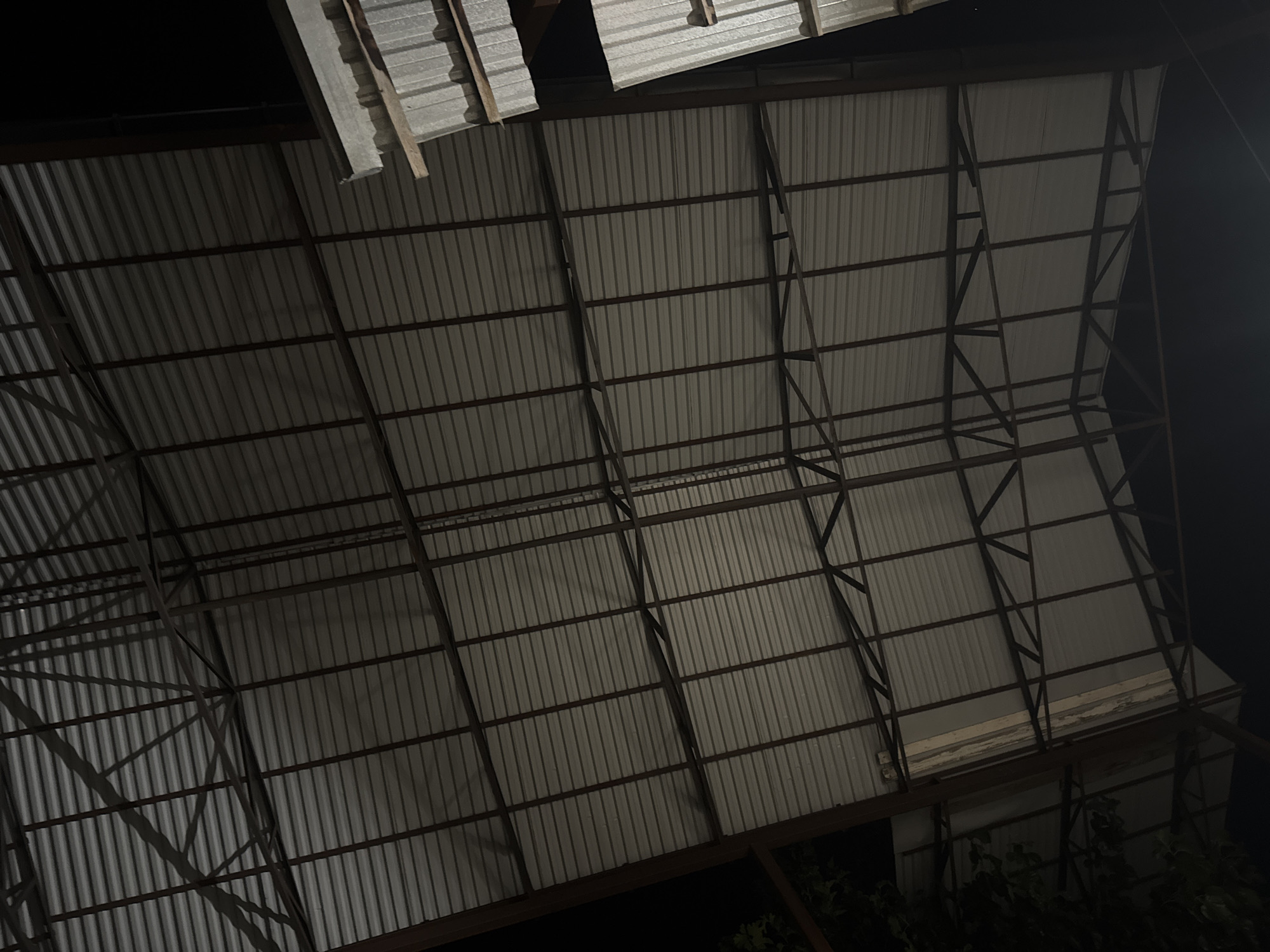 The image depicts the underside of a corrugated metal roof supported by a network of dark metal beams. The lighting suggests it is nighttime or the interior is dimly lit. The roof panels run horizontally across the frame, with vertical beams spaced at regular intervals. Diagonal bracing beams connect the vertical supports, creating a grid-like structure. On the right side of the image, a section of the roof appears to be constructed from larger, layered metal sheets, possibly insulation panels. Some foliage is visible on the far left, silhouetted against the darkness, suggesting the structure is outdoors. The scene is devoid of any people or discernible activity. The image provides a structural overview of a building's roof, likely a warehouse, industrial building, or open-air shelter.
