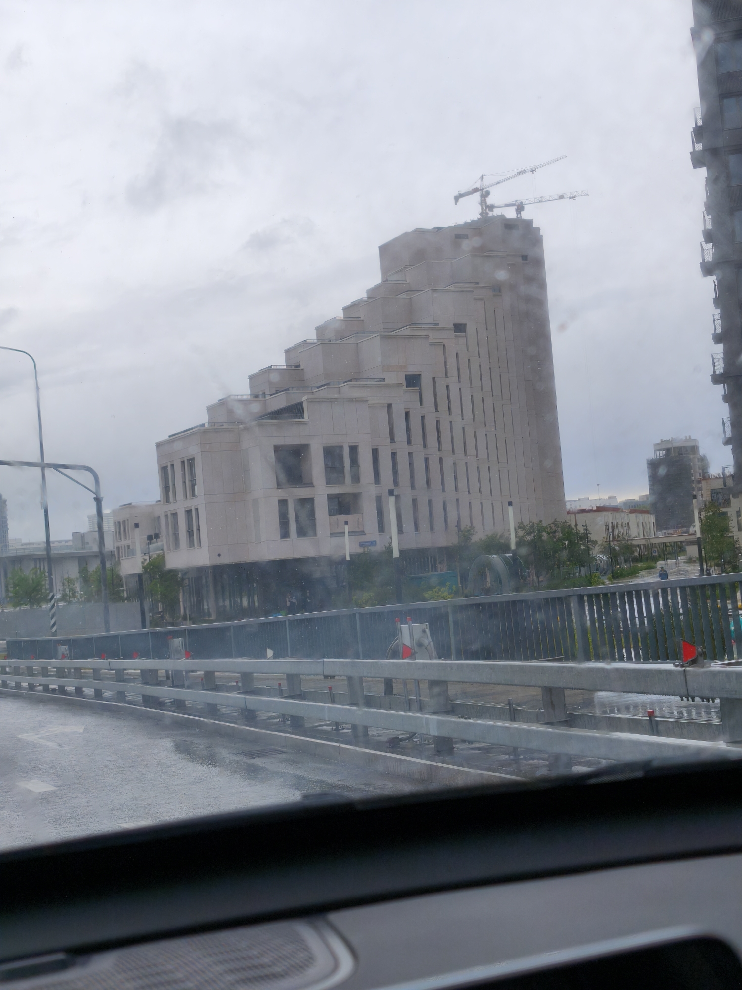 The image, taken from inside a vehicle, shows a partial view of the Khoroshevo-Mnevniki district in Moscow, Russia. A modern, tiered building under construction dominates the scene, with two construction cranes visible atop it. Another high-rise building is partially visible on the right side. The scene is viewed through a wet windshield, likely due to rain. A gray metal barrier is present along the road, indicating the location is likely a bridge or elevated roadway. The overall impression is one of urban development and ongoing construction in cloudy weather.