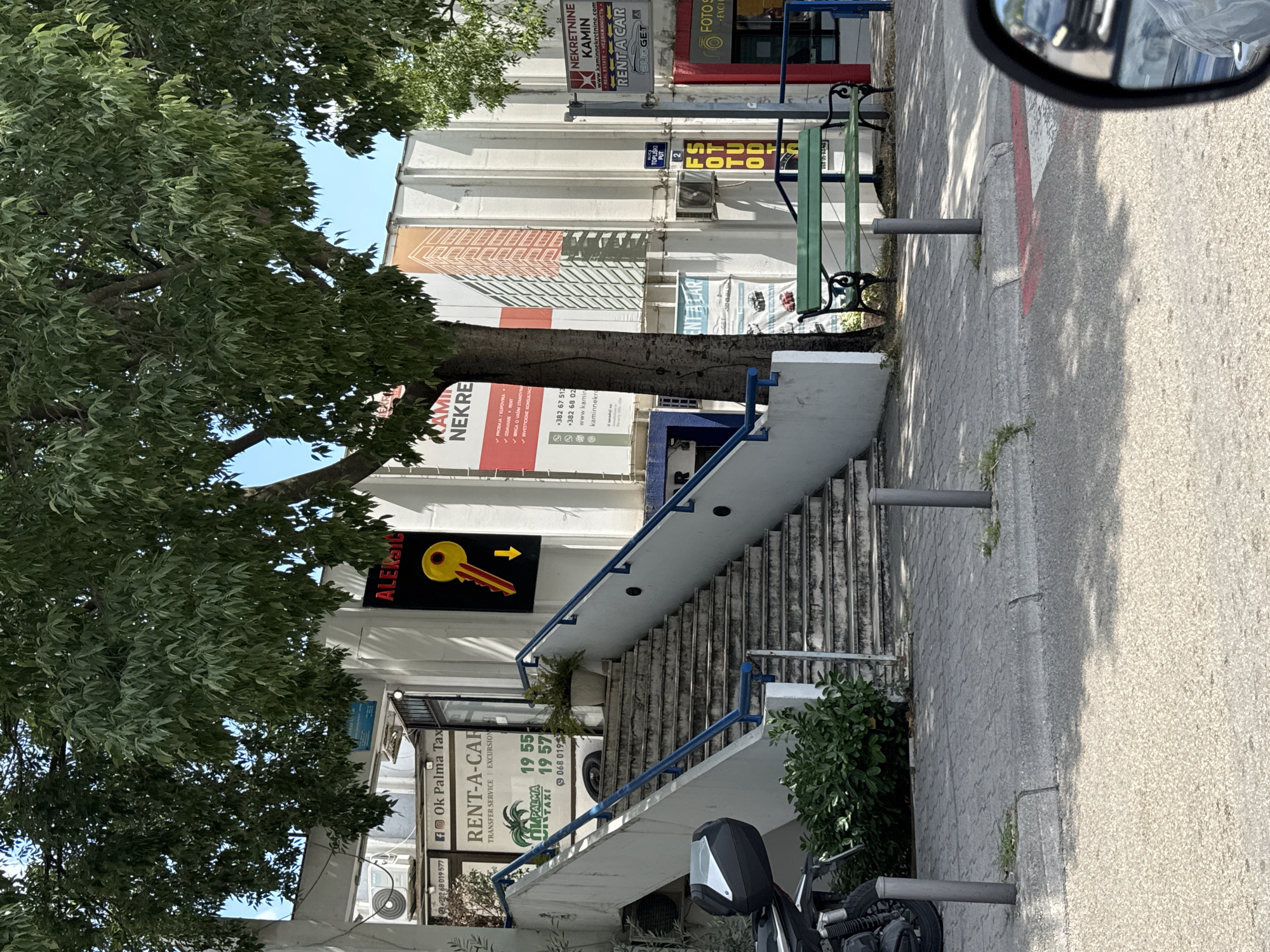 This is an image of a street scene in Budva, Montenegro, taken from a vehicle, as indicated by the reflection of the rearview mirror in the upper right corner. The image is oriented vertically.
The scene features a multi-story building with a white facade, adorned with various signs advertising businesses. Prominently displayed are signs for "NEKRETNINE KAMIN" (real estate), "RENT A CAR," and "FOTOS." There are also signs in Croatian, such as "AMIN NEKRE" and "PRODAJA/KUPOVINA" (selling/buying). Another sign indicates "Ok Palma Tax" with phone numbers and a website for "kaminnekretnine." Further down, a sign for "RENT-A-CAR" and "PALMA TAXI" with phone numbers and "TRANSFER SERVICE EXCURSION" is visible.
A large tree with lush green foliage is in the foreground, casting shadows on the building and the paved sidewalk. A set of outdoor stairs with blue railings leads up to the building. A green park bench is situated on the sidewalk. A motorcycle is parked at the bottom of the image. The time of day appears to be daytime, with bright sunlight creating distinct shadows. The weather seems to be clear and sunny. The overall impression is of a busy commercial street.