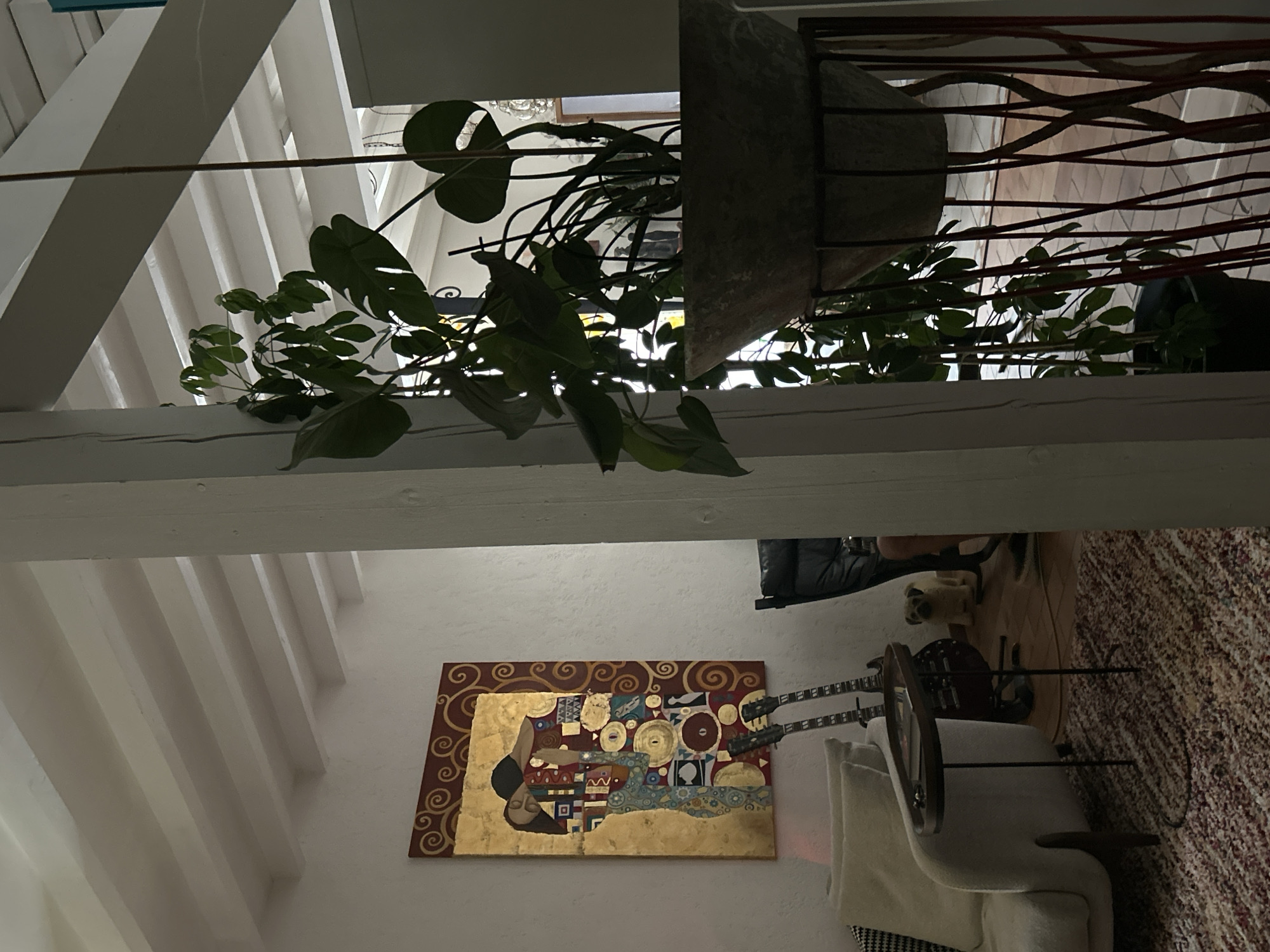 The image depicts an interior scene. On the left wall, a rectangular painting is displayed, featuring a depiction of two figures embracing, rendered in a style reminiscent of Gustav Klimt's "The Kiss," with prominent use of gold leaf and decorative patterns. Below the painting, two guitars are leaning against the wall. In the foreground, a beige sofa is partially visible, with a small, dark-topped side table next to it. A patterned rug covers the floor. To the right of the sofa, a person's legs are visible, suggesting a seated individual. A small, light-colored dog stands on the floor between the sofa and a black chair.
Dominating the right side of the image is a large potted plant with broad green leaves, housed in a light-colored, textured pot. The pot is supported by a dark, artistic stand with vertical elements. In the background, through an opening, a glimpse of another room is visible, with what appears to be a decorative chandelier and possibly more furniture.
The overall environment suggests a living space, possibly a home or a studio, with a blend of artistic and natural elements. The lighting is subdued, creating a warm and intimate atmosphere. The location is specified as Saint-Marcel-lès-Sauzet, France.