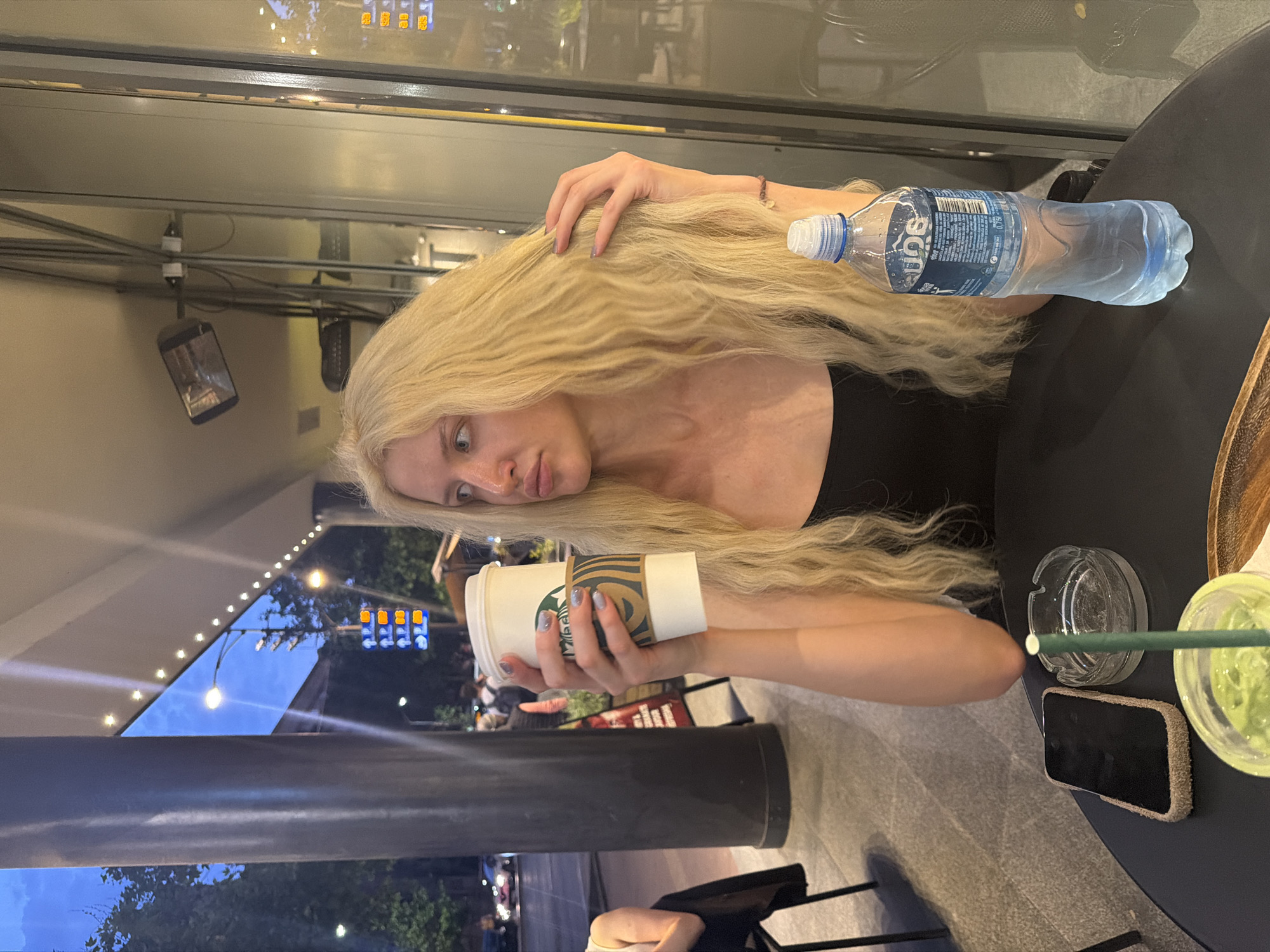 A woman with long blonde hair sits at a table in what appears to be an outdoor cafe in Vracar Urban Municipality, Serbia. She is holding a Starbucks coffee cup. A water bottle is on the table next to her hair. An ashtray, a drink, and a cell phone are also on the table. The background shows a cityscape and streetlights reflected in a window.