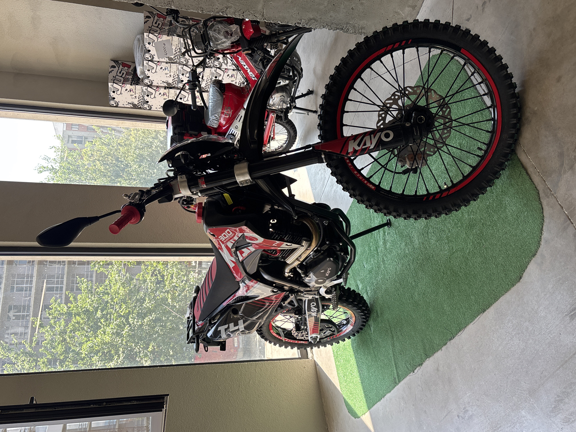 A black and red Kayo dirt bike is parked on artificial turf in front of a large window that looks out onto a tree-lined street and buildings in Tbilisi, Georgia. The dirt bike has a red seat, red accents on the wheels, and various decals. In the background, another red motorcycle is visible, along with stacked boxes labeled "JUST1". The scene appears to be an indoor showroom or garage.
