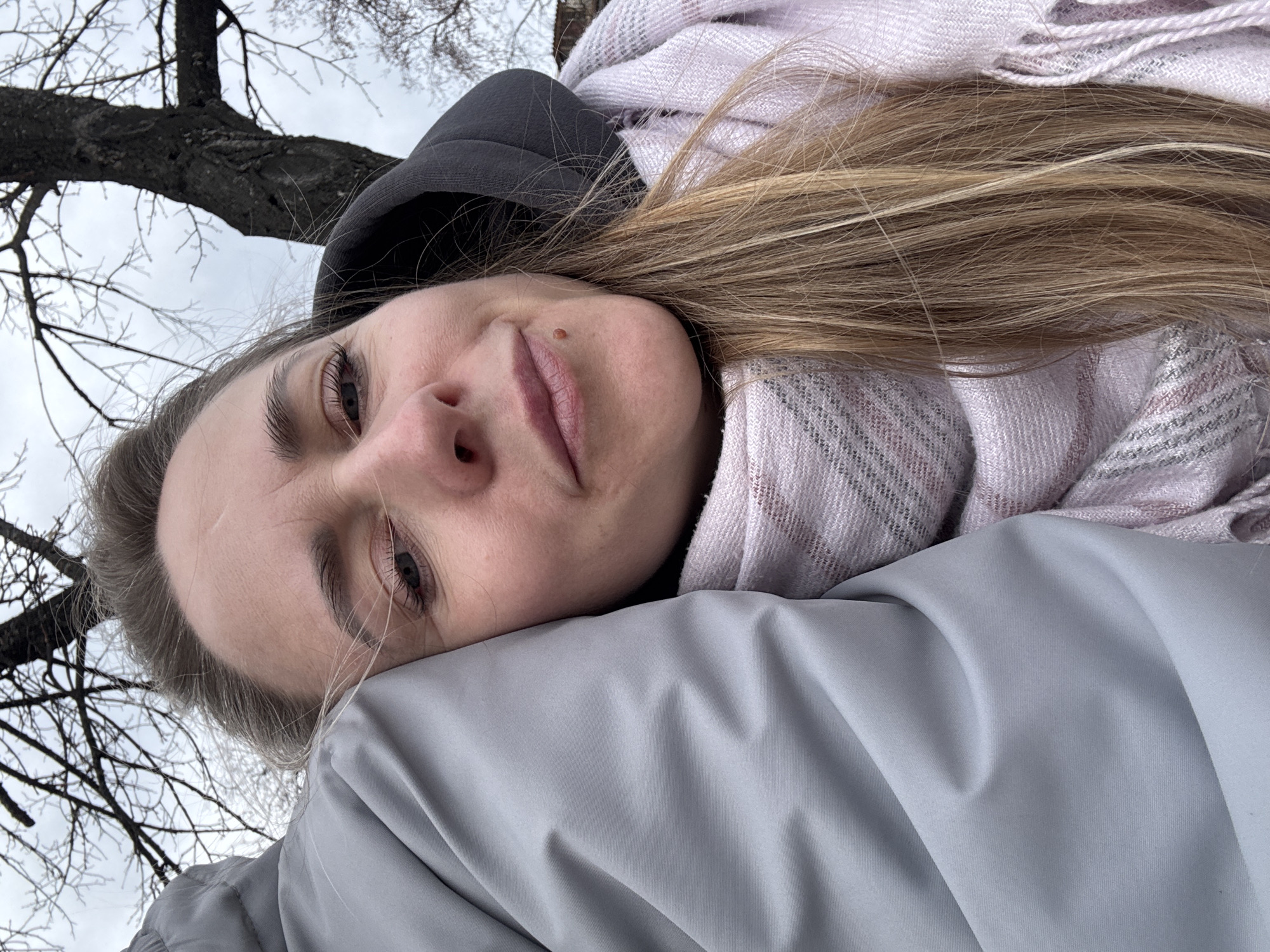 A woman lies with her head resting on a light gray padded surface, possibly a jacket or coat. She is wearing a dark gray hooded sweatshirt and a light pink and white patterned scarf is wrapped around her neck. Her blonde hair is splayed across the scarf and padding. She looks directly at the camera with a neutral expression. In the upper left corner, bare tree branches are visible against an overcast sky. The image was likely taken in an outdoor environment on a day with limited sunlight. The location is mentioned as Chaikovsky, Russia, but there are no specific location indicators in the photo.