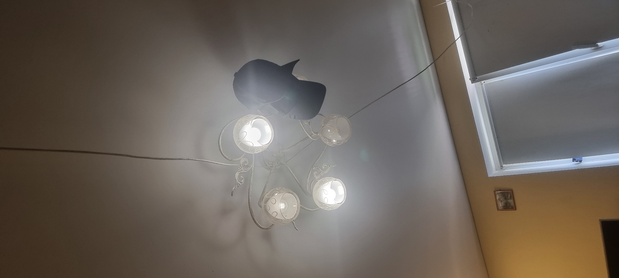 A black baseball cap is draped over a white, ornate ceiling light fixture with five glass globes. The cap hangs from the fixture, obscuring one of the lights. The light fixture is suspended from a thin cord that runs vertically along the ceiling. In the background, a wall painted a light yellow color is visible, with a small framed picture hanging on it. To the right, a window with a closed white blind is partially visible. The overall impression is an indoor residential setting.