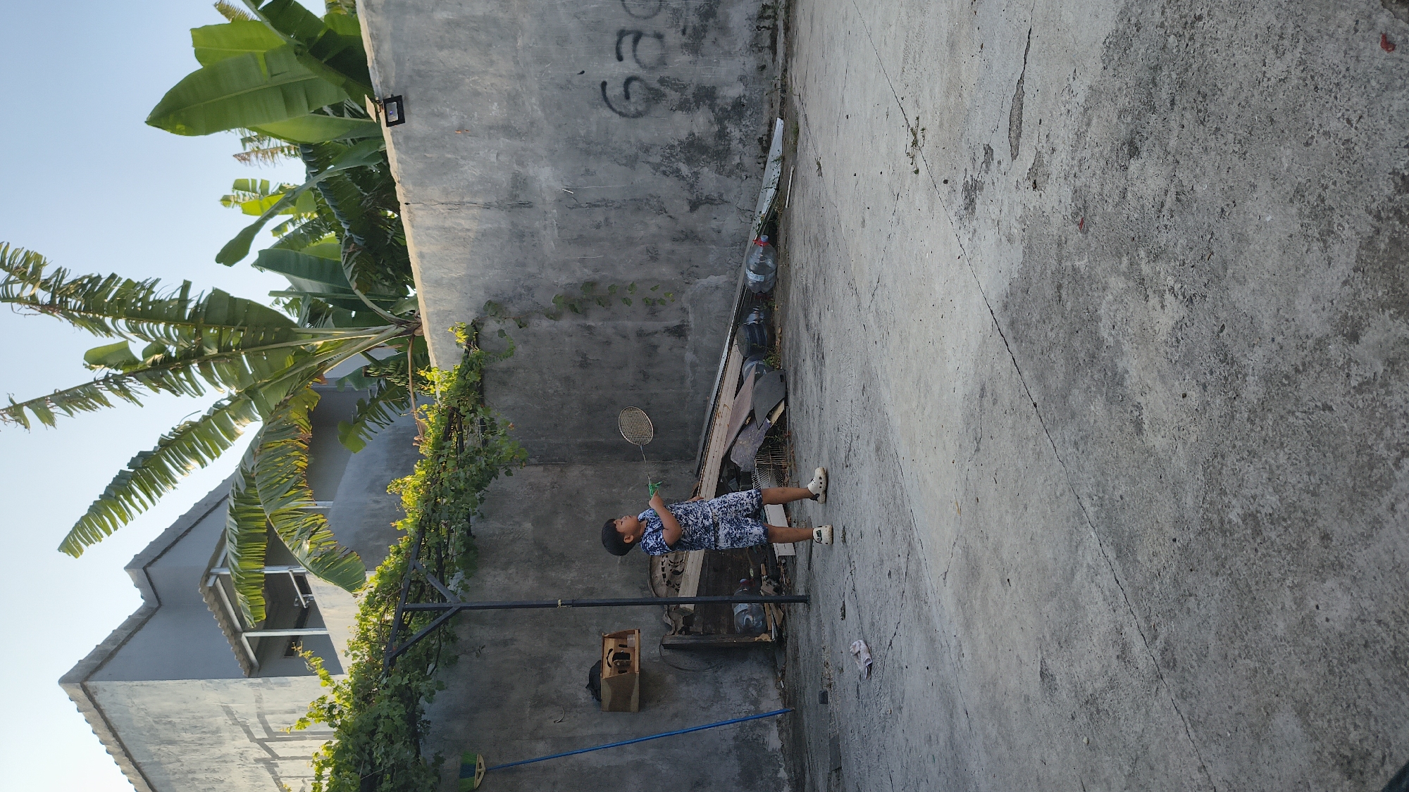 In Setu Wetan, Indonesia, a young boy stands on a concrete surface, engaged in playing badminton. He is wearing a blue and white patterned t-shirt and shorts, with white clogs on his feet. The boy holds a badminton racket in his right hand, mid-swing. The background features a rough, gray concrete wall with some graffiti. Behind the wall, banana trees with large green leaves are visible, along with a portion of a building. The ground is a cracked concrete patio, with discarded materials like plastic bottles and wood scraps piled near the wall. A blue broom leans against the wall to the left of the boy. The scene is illuminated by natural sunlight, suggesting it is daytime.