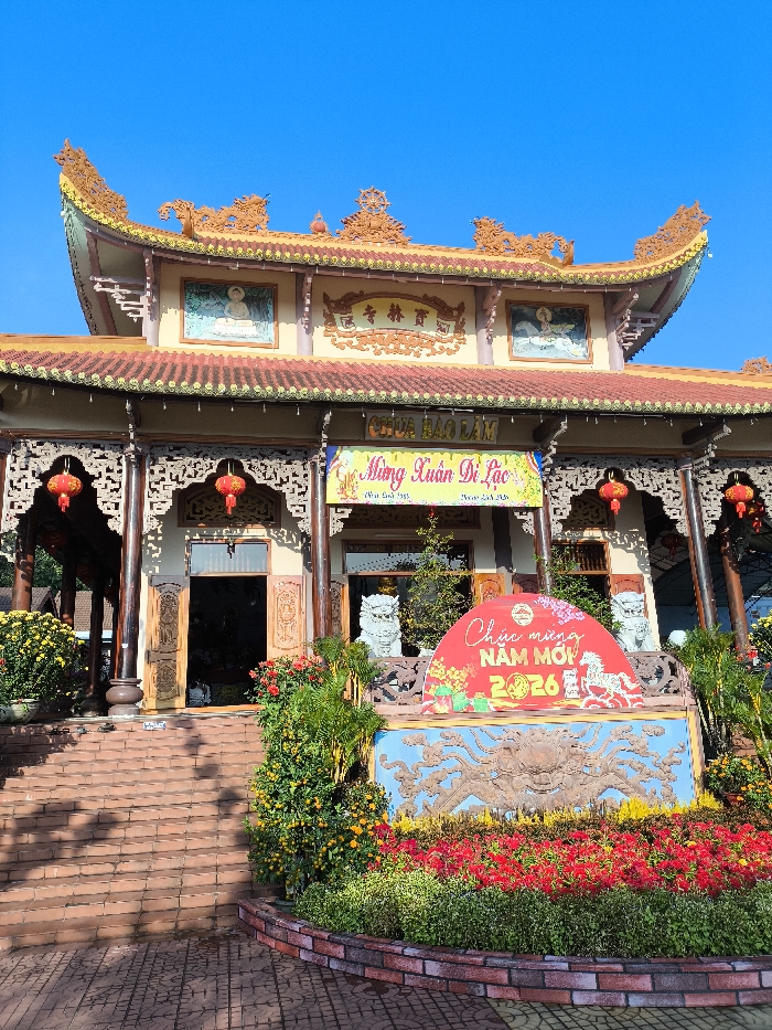 The image displays the exterior of Chùa Bảo Lâm (Bao Lam Pagoda) in Thành phố Tuy Hòa, Vietnam. The building exhibits traditional Vietnamese Buddhist architecture with a multi-tiered, ornate roof featuring red tiles and intricate gold decorations. The facade includes carved wooden elements, pillars, and two pictorial plaques depicting Buddhist figures on the upper level.
A yellow banner across the building's front displays "CHÙA BẢO LÂM," with another banner below it reading "Mừng Xuân Di Lặc." Red lanterns are suspended near the main entrance. A wide set of red-brown brick steps leads up to the pagoda's entrance. The area surrounding the steps and the building's base is adorned with various potted plants, including yellow chrysanthemums and kumquat trees, along with planted red and yellow flowers. Two white, stylized lion statues flank the central entrance.
In the foreground, a prominent New Year display is situated. It consists of a large blue relief depicting a crab-like motif, topped by a semi-circular red sign. This sign is inscribed with "Chúc mừng NĂM MỚI 2026" (Happy New Year 2026) and features a graphic of a white horse. The scene is illuminated by natural light under a clear blue sky. No individuals are visible in the image.
