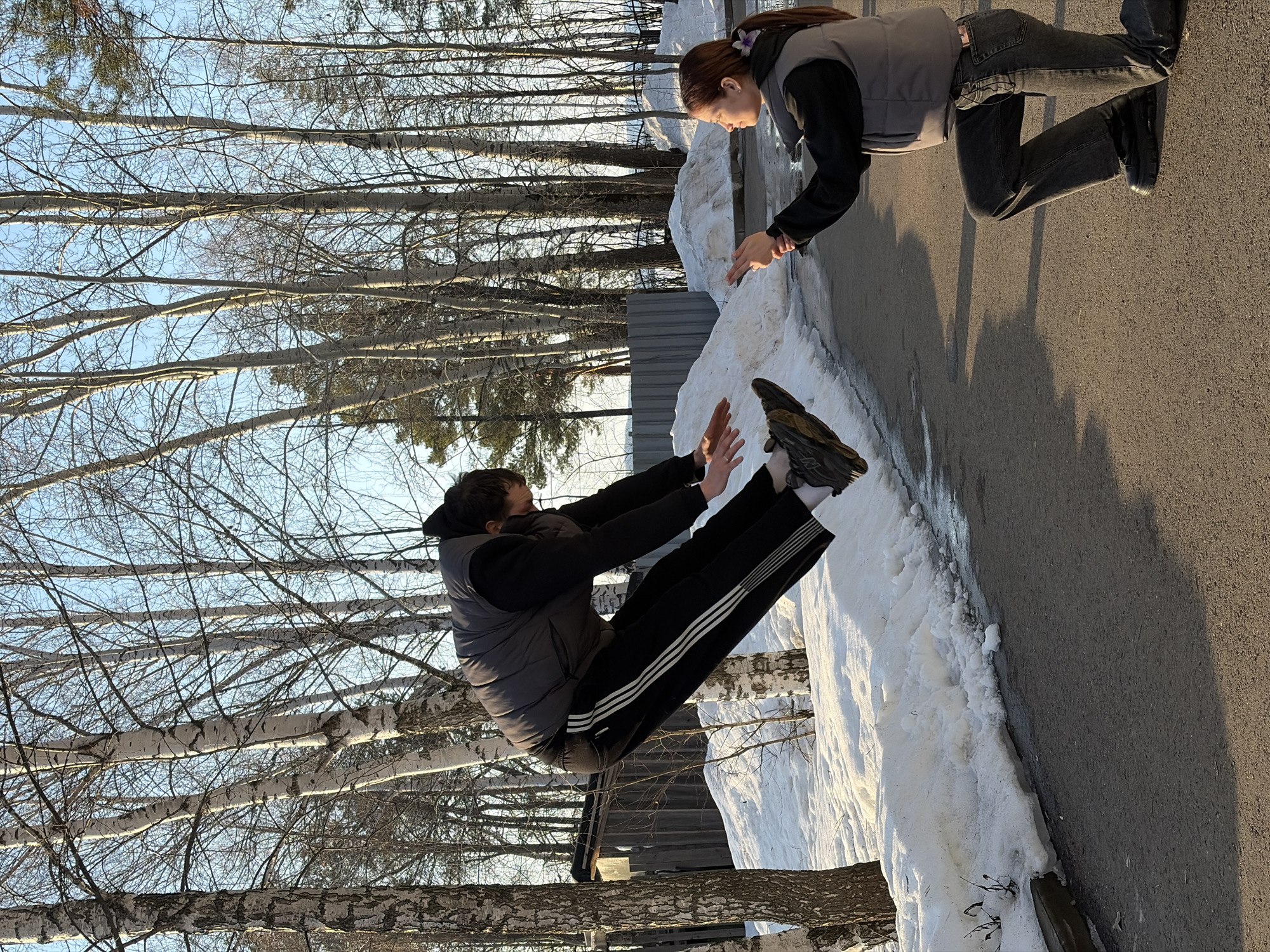 Two people are present in an outdoor scene. One person, wearing a gray vest and black pants with white stripes, is positioned in mid-air, hands raised. The other person, also in a gray vest and dark clothing, stands at the edge of a snow-covered area, reaching out toward the first person. The location appears to be near a paved surface adjacent to a snow bank. A line of birch trees is visible in the background against a blue sky. The scene is located in the city of Chaikovsky, Russia.