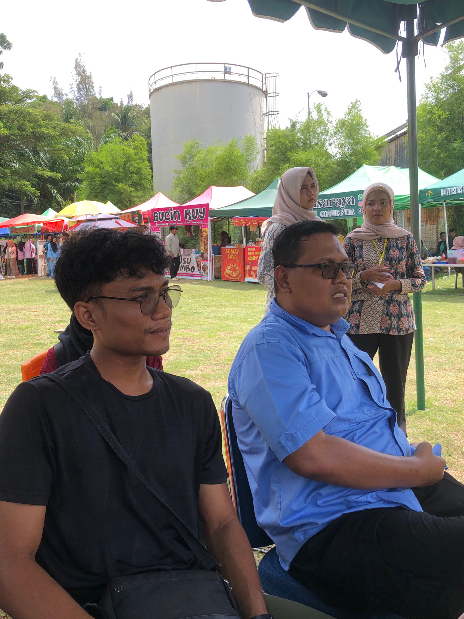The image depicts an outdoor event, likely a bazaar or festival, set in a grassy area with trees in the background and a large cylindrical water tank. In the foreground, two men are seated, facing towards the right of the frame. The man on the left is wearing a black t-shirt and glasses. The man on the right is wearing a blue button-down shirt and glasses. Behind them, several stalls with colorful canopies are set up, with people milling about. Some stalls have signage, including one with the text "BUCIN KUY" and another that appears to be for "SARAYA COFFEE & TEA." Further in the background, a few individuals are visible, including two women wearing hijabs. The overall atmosphere suggests a casual, community gathering. The presence of the water tank and the general environment are consistent with locations in Lhokseumawe, Indonesia.
