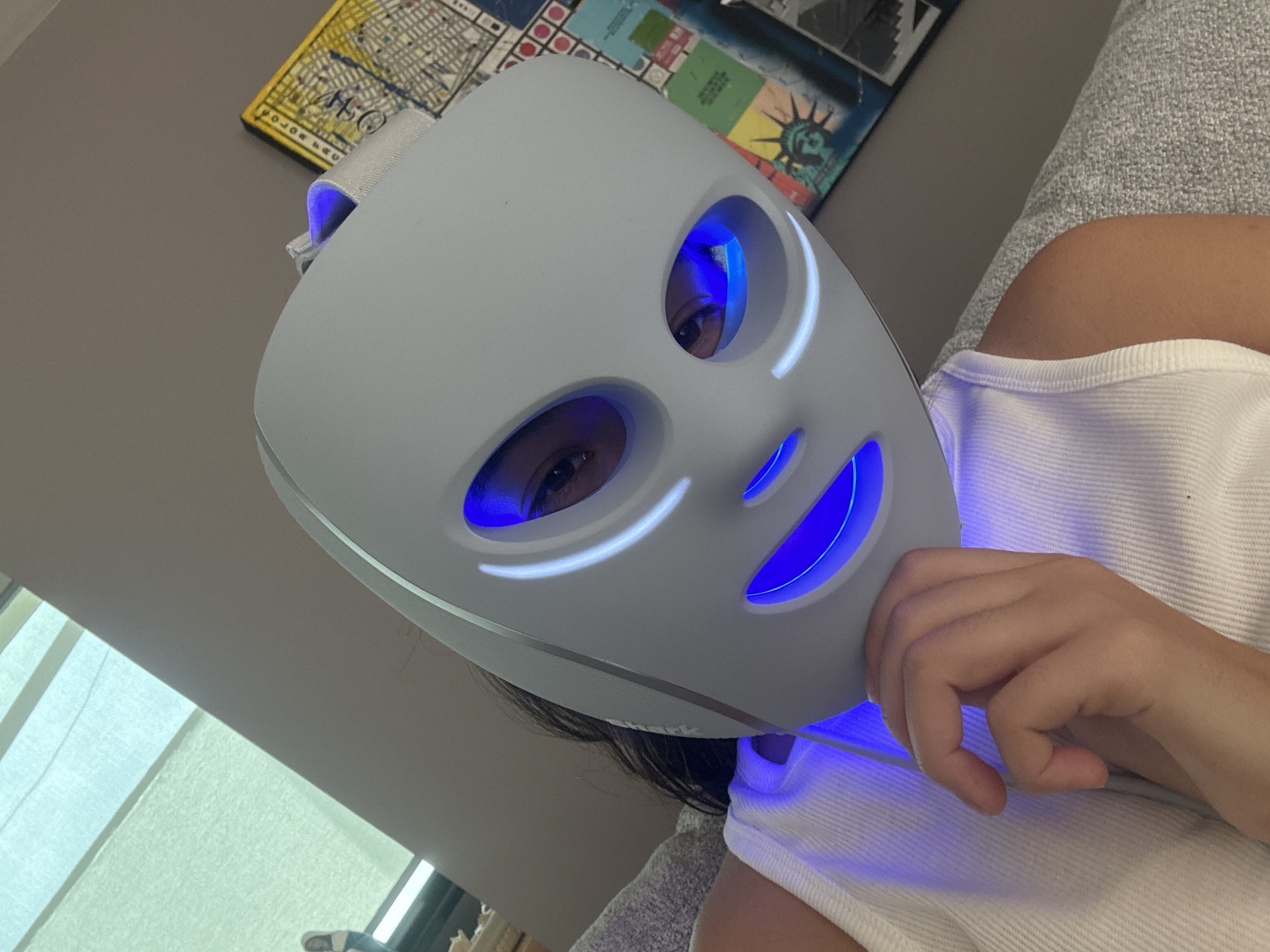 A person is wearing a white LED face mask that emits blue light. The person is wearing a white tank top and lying on a gray textured surface, possibly a couch. The eyes of the person are visible through the eye openings of the mask. In the background, there is a wall with artwork on it, including a depiction of the Statue of Liberty. The location is San Bernardino Tlaxcalancingo, Mexico.