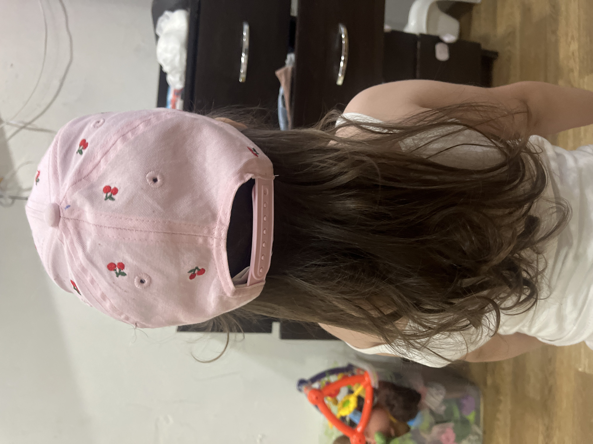 The image shows the back of a young child wearing a pink baseball cap with embroidered cherries. The child has long, brown hair that is visible from under the cap and falls down their back. They are wearing a white t-shirt. The child appears to be indoors, with a dark dresser with shiny handles in the background. A white object, possibly a changing table or a stool, is also visible in the background, along with a colorful toy in the lower right corner of the image. The scene is dimly lit, suggesting it might be daytime or evening. The overall impression is a casual, domestic setting, likely a home. No text is visible in the image.