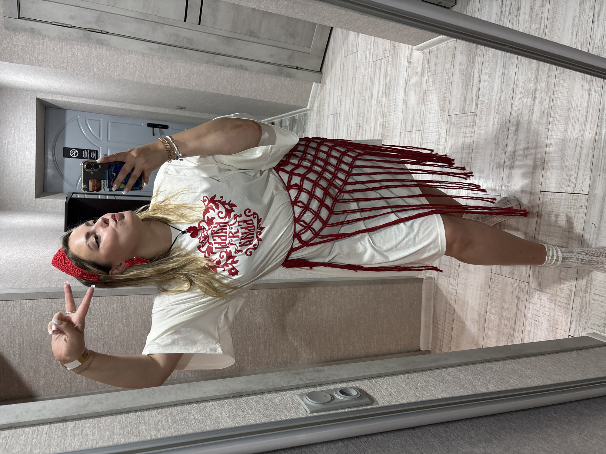 A woman is taking a mirror selfie indoors. She is wearing a white t-shirt dress with red embellishments on the front, a red macrame-style belt with fringe, white fishnet socks, and white sneakers. She has on red heart-shaped earrings and a red headband. She is holding her left hand up in a peace sign. The background appears to be a hallway with light-colored walls and light wooden flooring. There is a door visible behind her to the right. The location is not specified as Kungur, Russia.