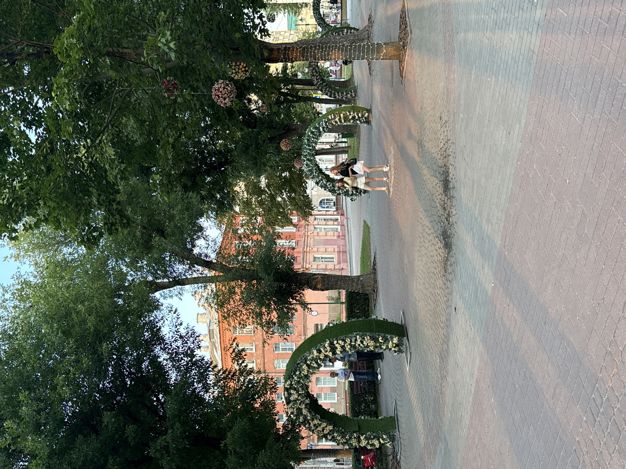 The image depicts a scene in the city of Mahilyow, Belarus, likely in a park or public square, characterized by decorative floral arches and lush trees. In the foreground, a paved walkway with a repeating brick pattern runs across the frame, with shadows cast by the surrounding trees indicating it is late afternoon or early evening.
Several large, arched structures, covered in artificial greenery and white and yellow flowers, are positioned along the walkway. The first arch, closer to the viewer, has two men standing near it. One man, wearing a white t-shirt and blue jeans, stands facing left, while another man, in a denim jacket and dark pants, stands to his right, looking at the first man. Behind them, a bench is visible.
Further down the walkway, two young women are walking towards the right. The woman on the left wears a light yellow top and white shorts, while the woman on the right wears a black top and a white skirt. They appear to be conversing or walking together.
The background features a large, red brick building with multiple windows, suggesting it is a historical or institutional structure. Mature trees with dense green foliage are scattered throughout the scene, some with decorative spherical floral arrangements hanging from their branches. One tree, to the right of the frame, is wrapped with string lights. The overall atmosphere is peaceful and suggests a well-maintained public space.