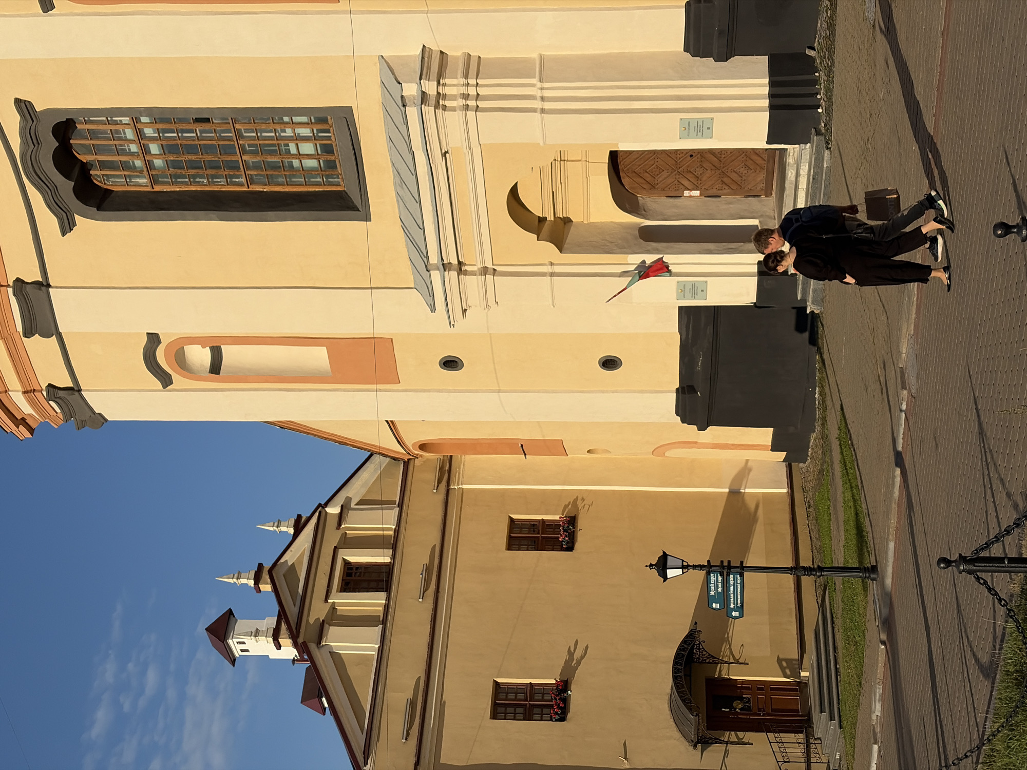 A young couple walks in front of a yellow and cream colored building with a dark gray trim. The building has a large, arched doorway with a wooden door and a flag hanging to the left of the entrance. The couple is walking from left to right, with the man carrying a shopping bag. The woman is wearing a black dress and the man is wearing a dark blue sweater and gray pants. The building is bathed in sunlight, casting long shadows on the ground. Behind the couple, to the left, is another yellow building with a pitched roof and several windows. There is a lamppost with directional signs in front of the building on the left. The scene takes place on a paved street. This is a view of the Holy Spirit Cathedral in Minsk, Belarus.