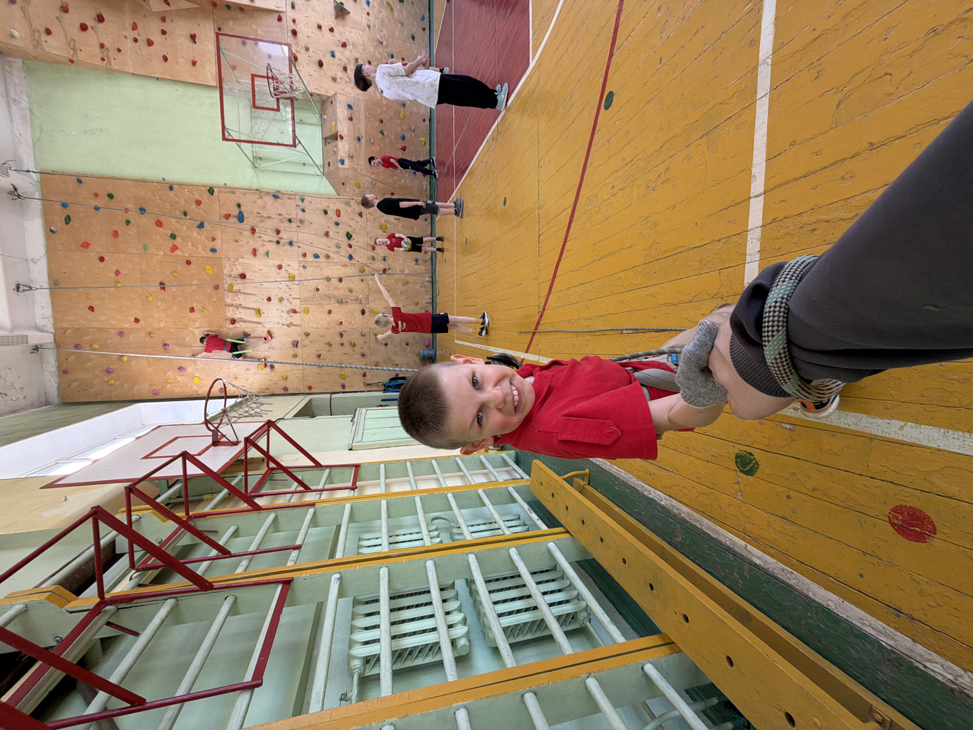 The image shows a young boy in a red shirt, harnessed, smiling at the camera, and being held by a person in a grey sweatshirt. Behind them is a gymnasium setting with a climbing wall featuring colored holds and multiple people climbing or preparing to climb. Basketball hoops are present, and a yellow wooden floor with markings is visible. The gymnasium has radiator-like heaters protected by metal bars. According to information given in the prompt, the gymnasium may be located in Izhevsk, Russia.