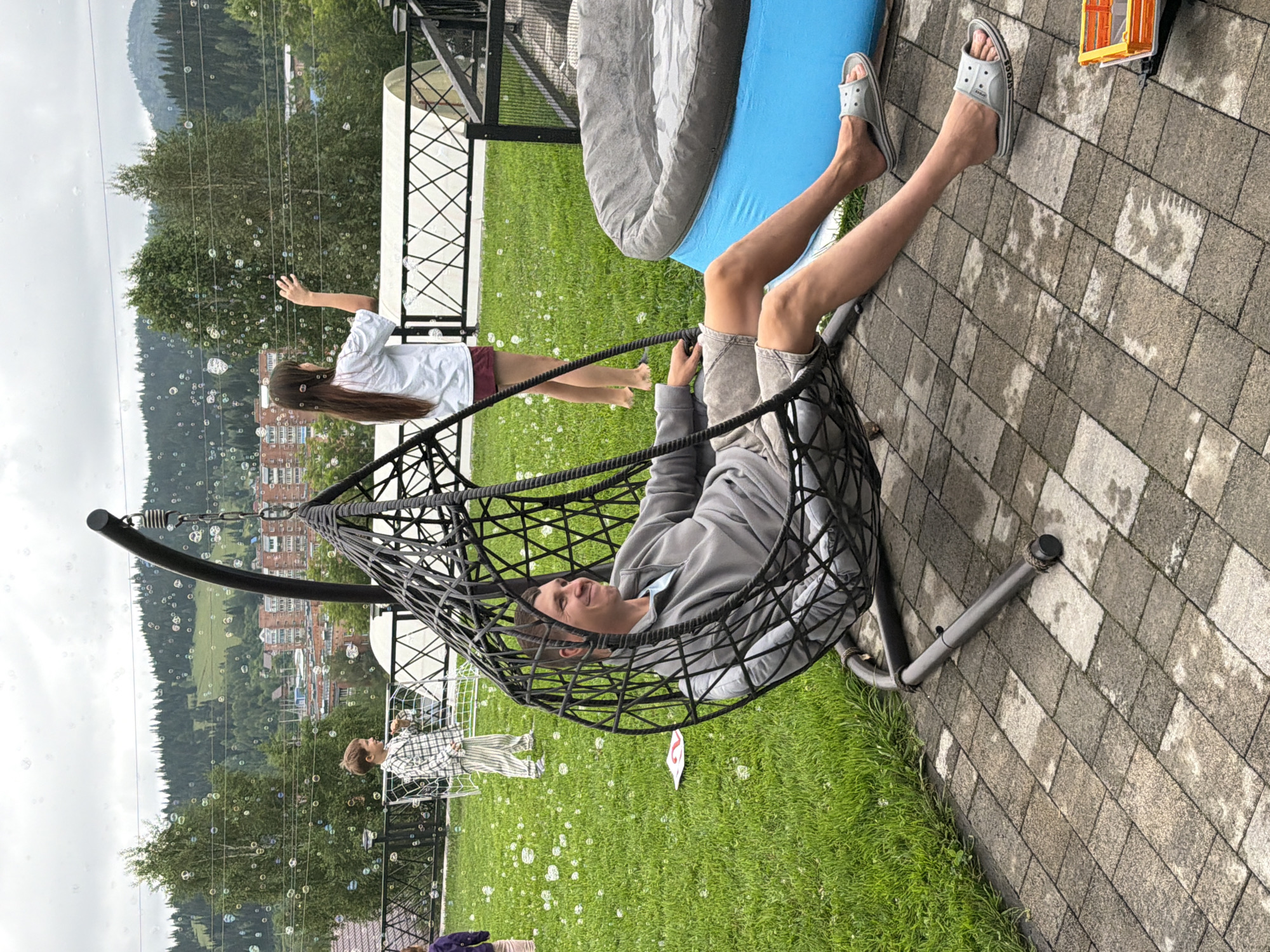 A man is sitting in a hanging chair in a backyard. The man is wearing a gray sweatshirt, gray shorts, and gray sandals. He is smiling and looking to the right. There are bubbles in the air. In the background, there is a girl standing and reaching her hand up towards the bubbles. There is also a boy standing in a soccer net. There is a blue inflatable pool in the background. The backyard is surrounded by trees and mountains. The city of Tashtagol, Russia, is visible in the distance.